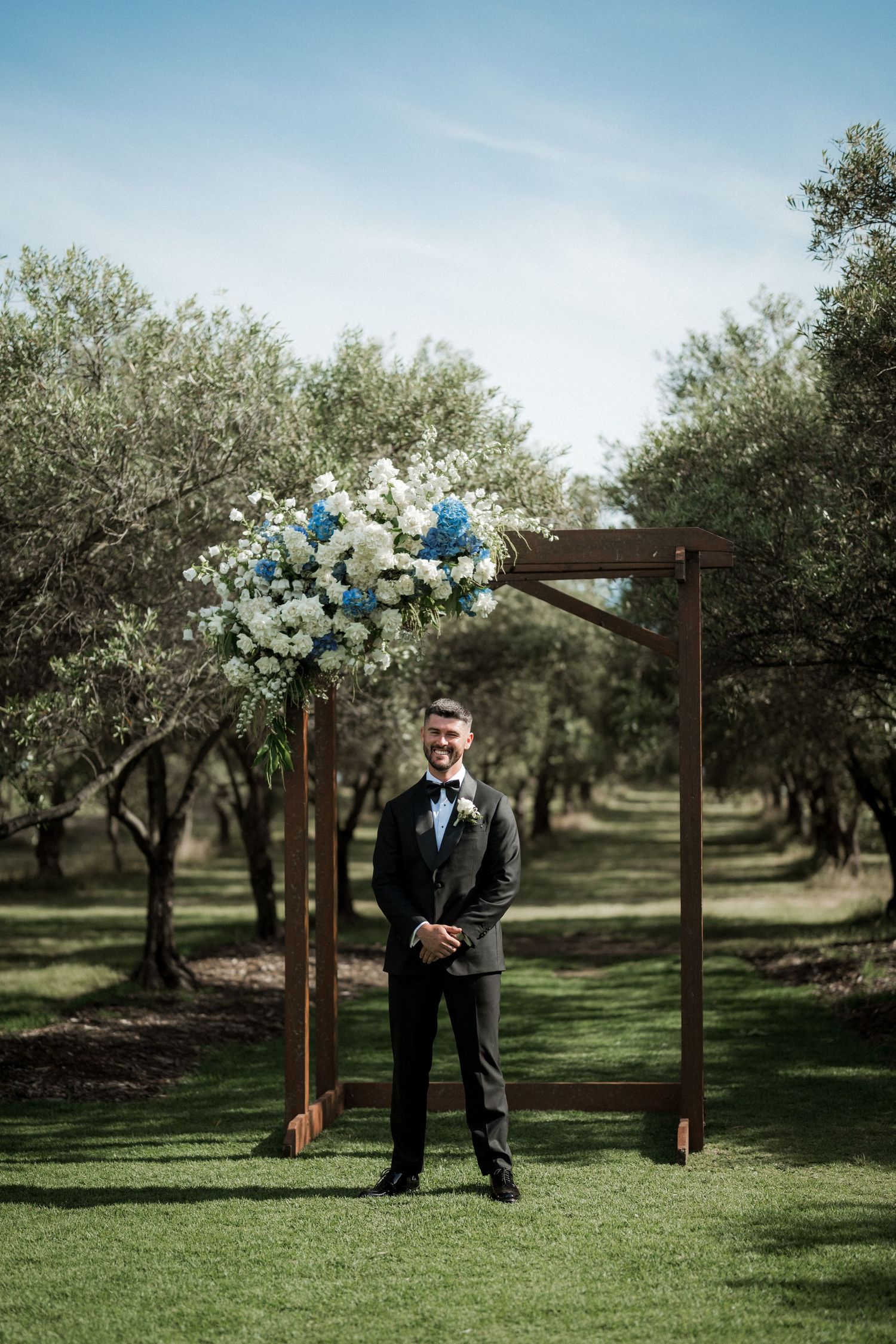 Rustic wooden wedding arch decorated with blue and white flowers in an olive grove.