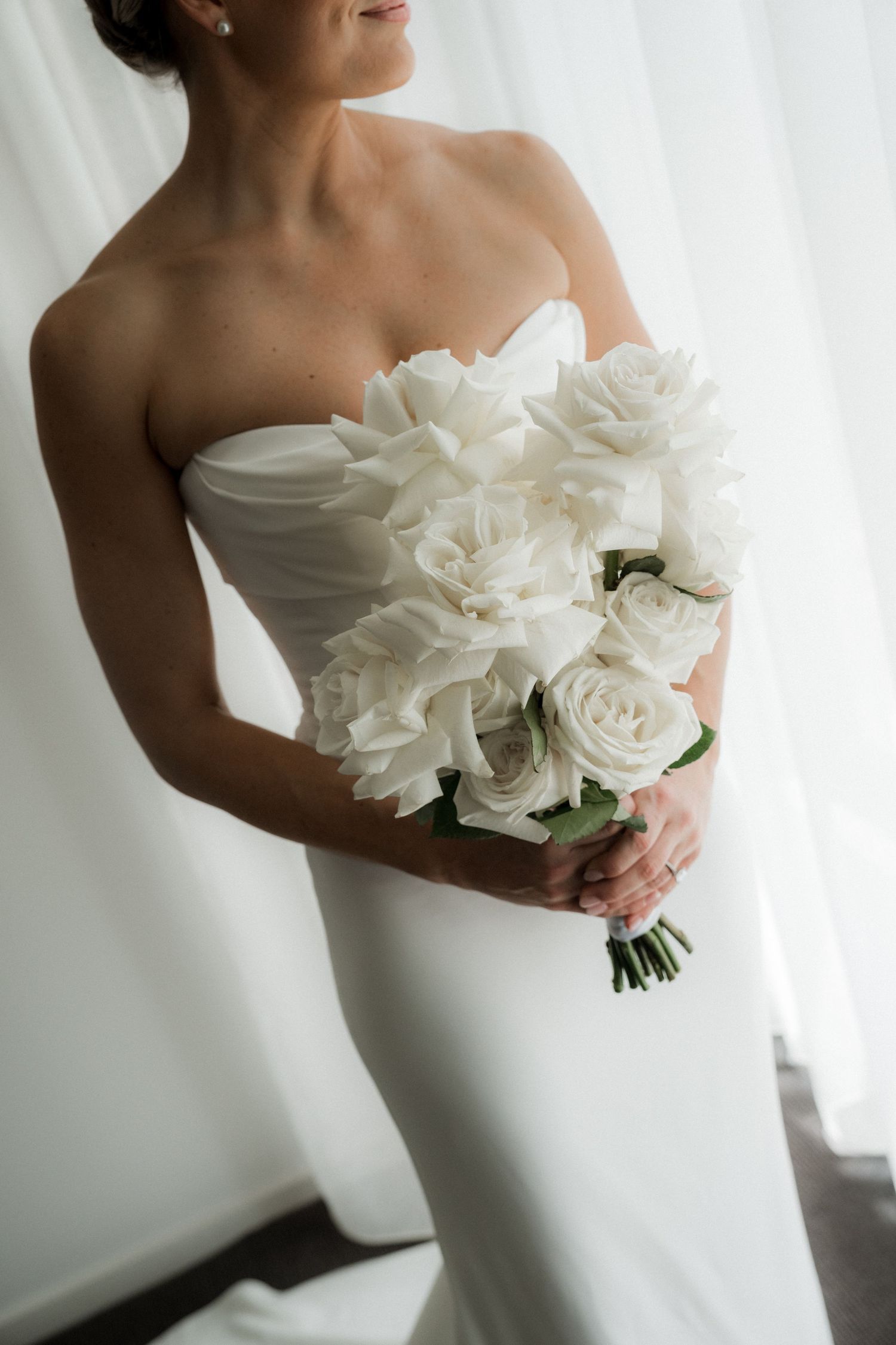 A bride in a white strapless wedding dress holds an elegant bouquet of white roses against a light background.