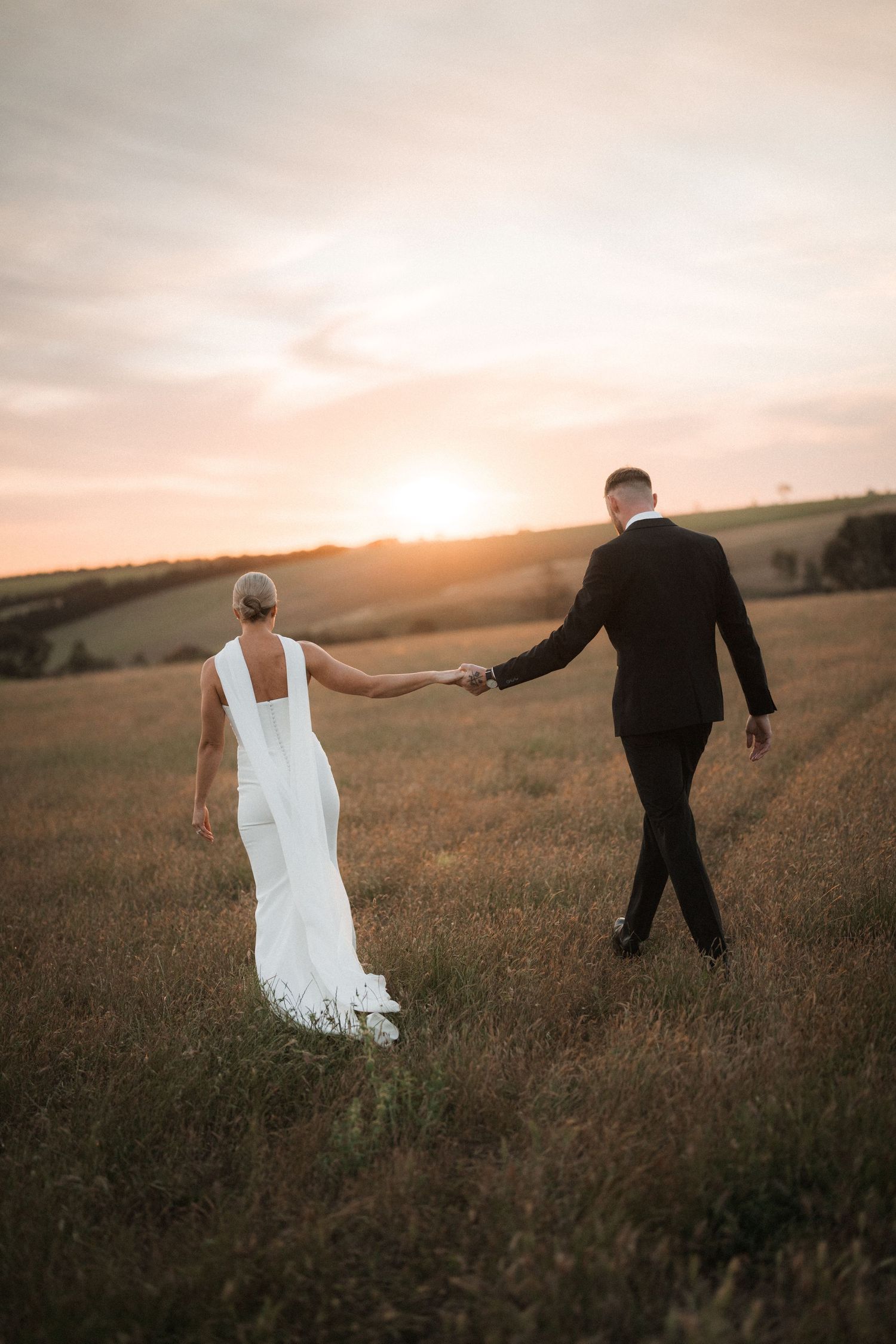 Couple walking hand in hand through field at sunset in wedding attire.