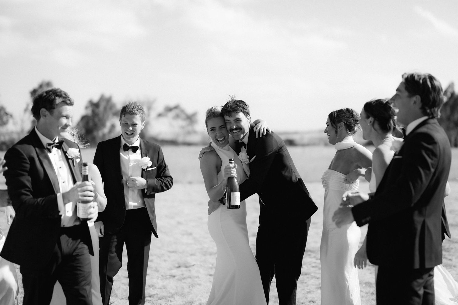Wedding party in formal attire sharing laughs during an outdoor celebration.