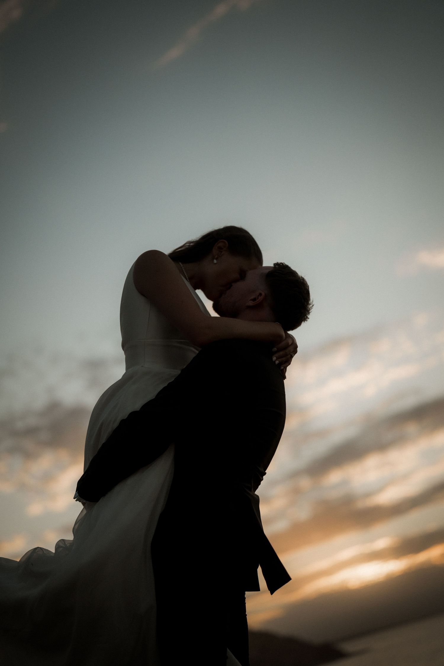 Silhouetted couple embracing against dramatic sunset sky during wedding photoshoot.