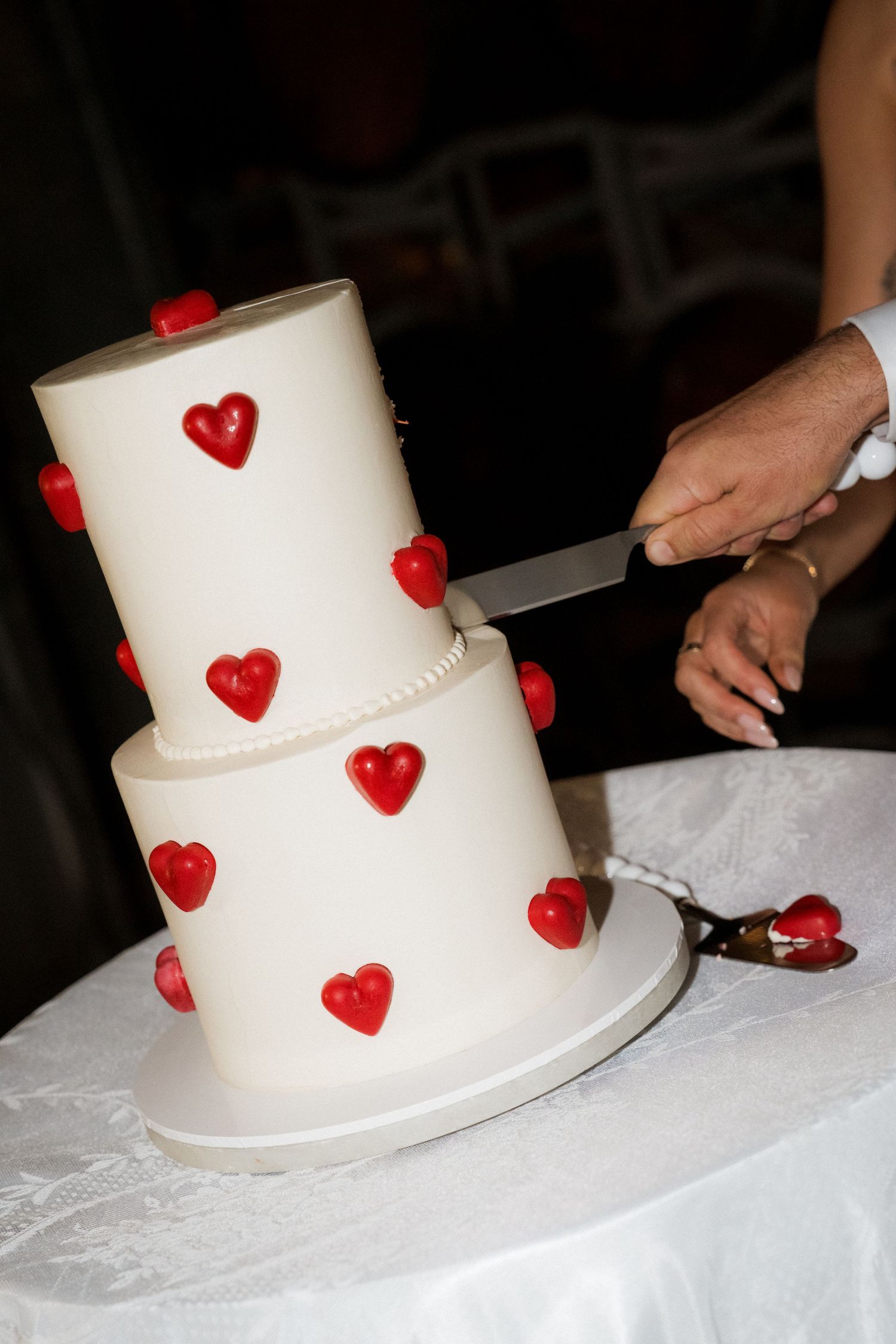 White tiered wedding cake decorated with small red heart details being cut.