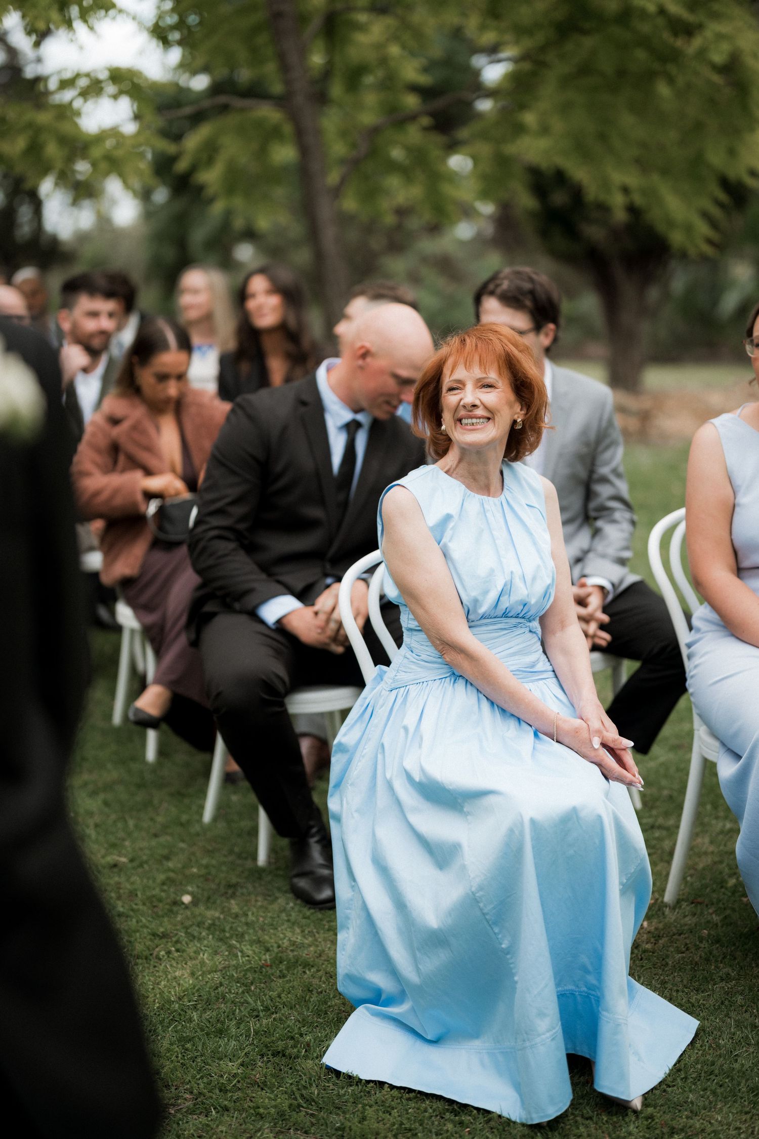 Wedding guests in light blue dresses seated on white chairs during an outdoor garden ceremony.