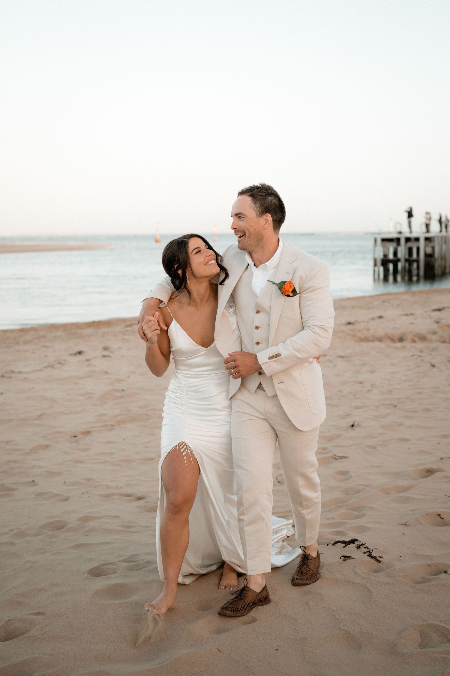 A couple in white attire shares romantic moments on a sandy beach at sunset near a wooden pier.