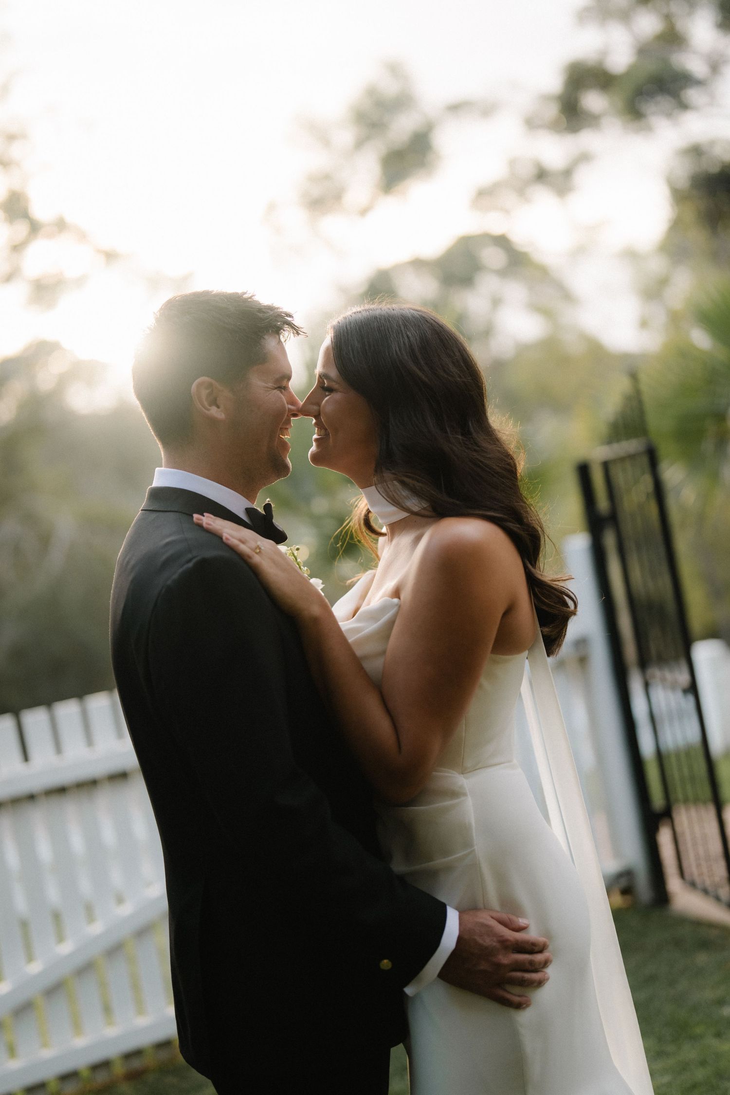 Romantic backlit couple embracing during golden hour with white picket fence in background.