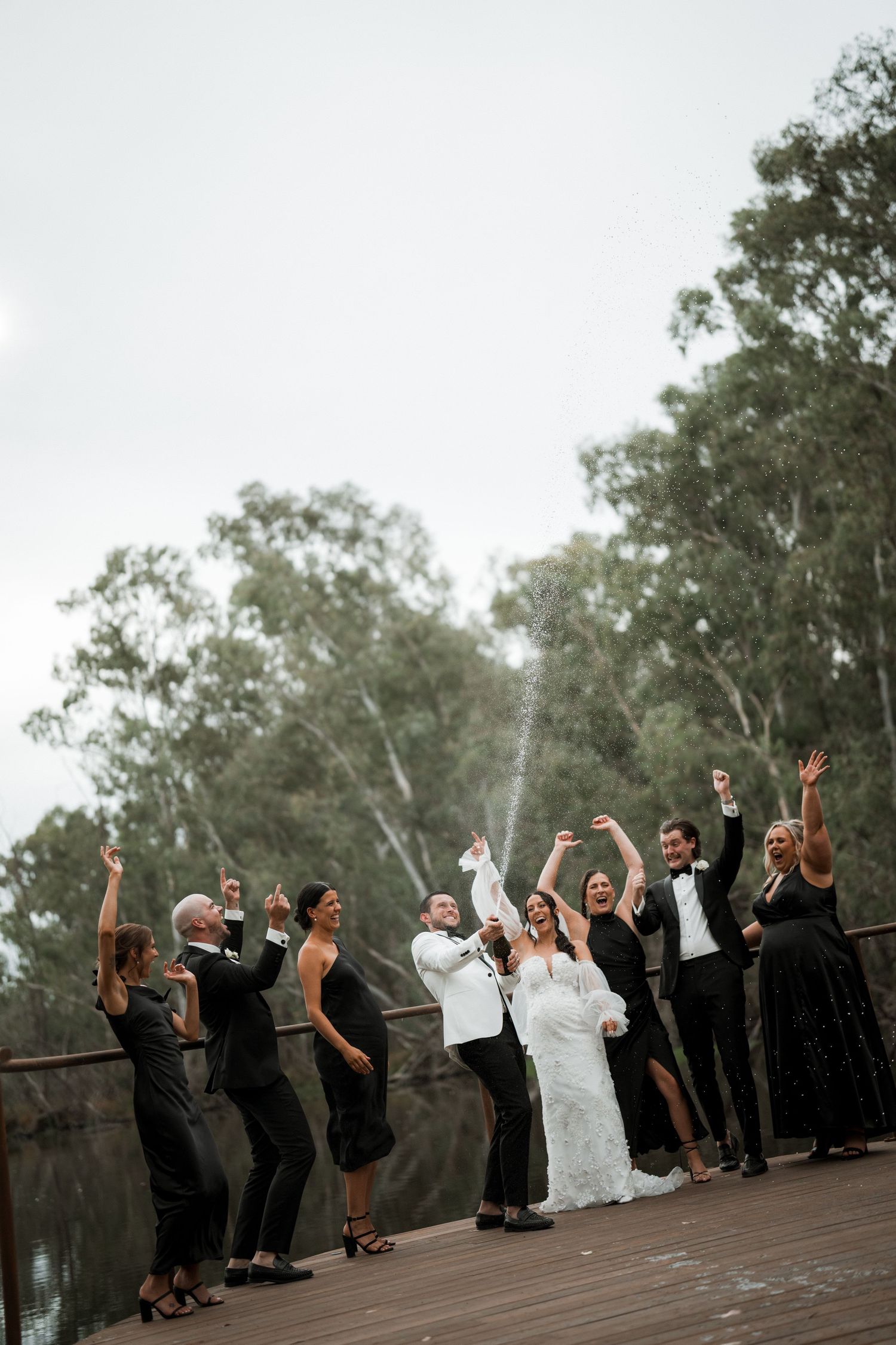 Wedding party celebrating with raised arms on outdoor deck against tree backdrop.