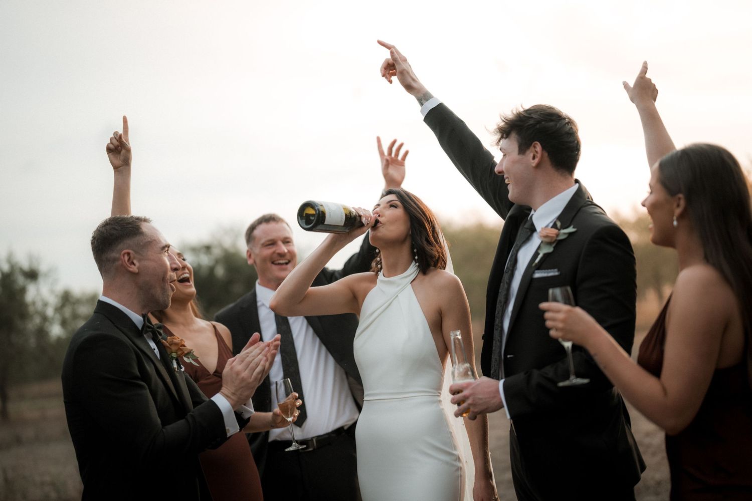 A bride and wedding guests celebrate outdoors with champagne and wine glasses while pointing upward at sunset.