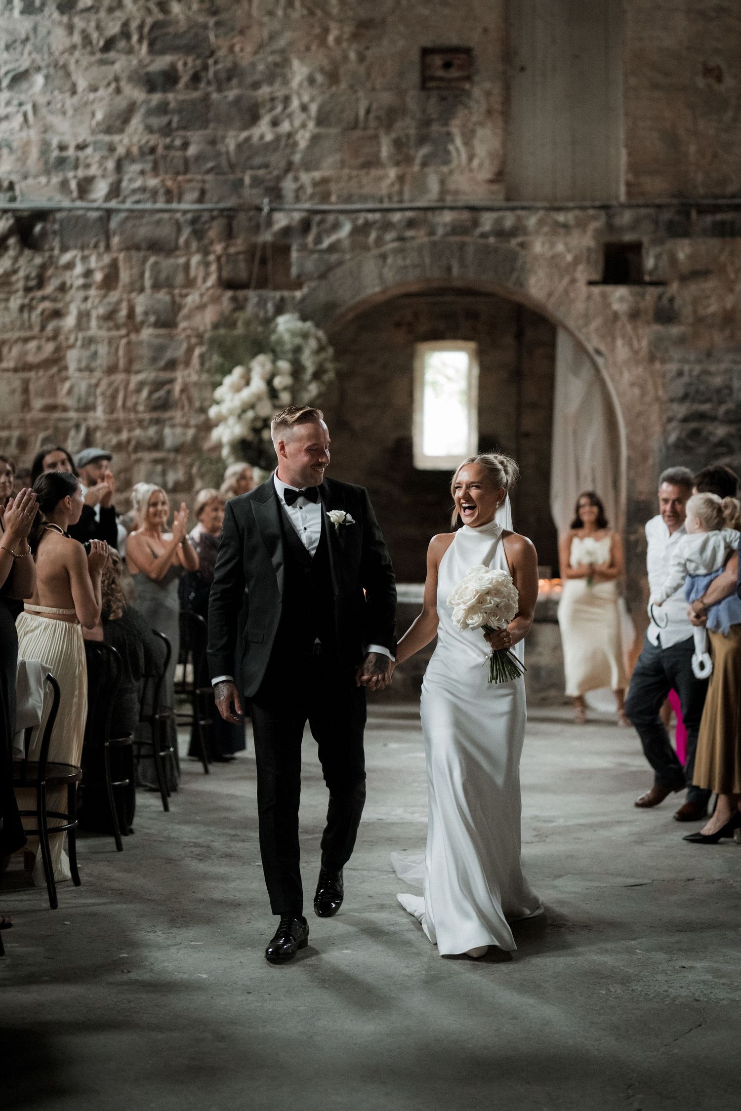 A bride and groom walk down the aisle together inside a rustic stone church building as guests watch from the sides.
