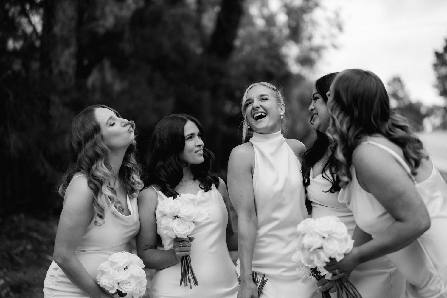 Bridesmaids in white dresses share a candid laughing moment while holding bouquets in black and white photography.