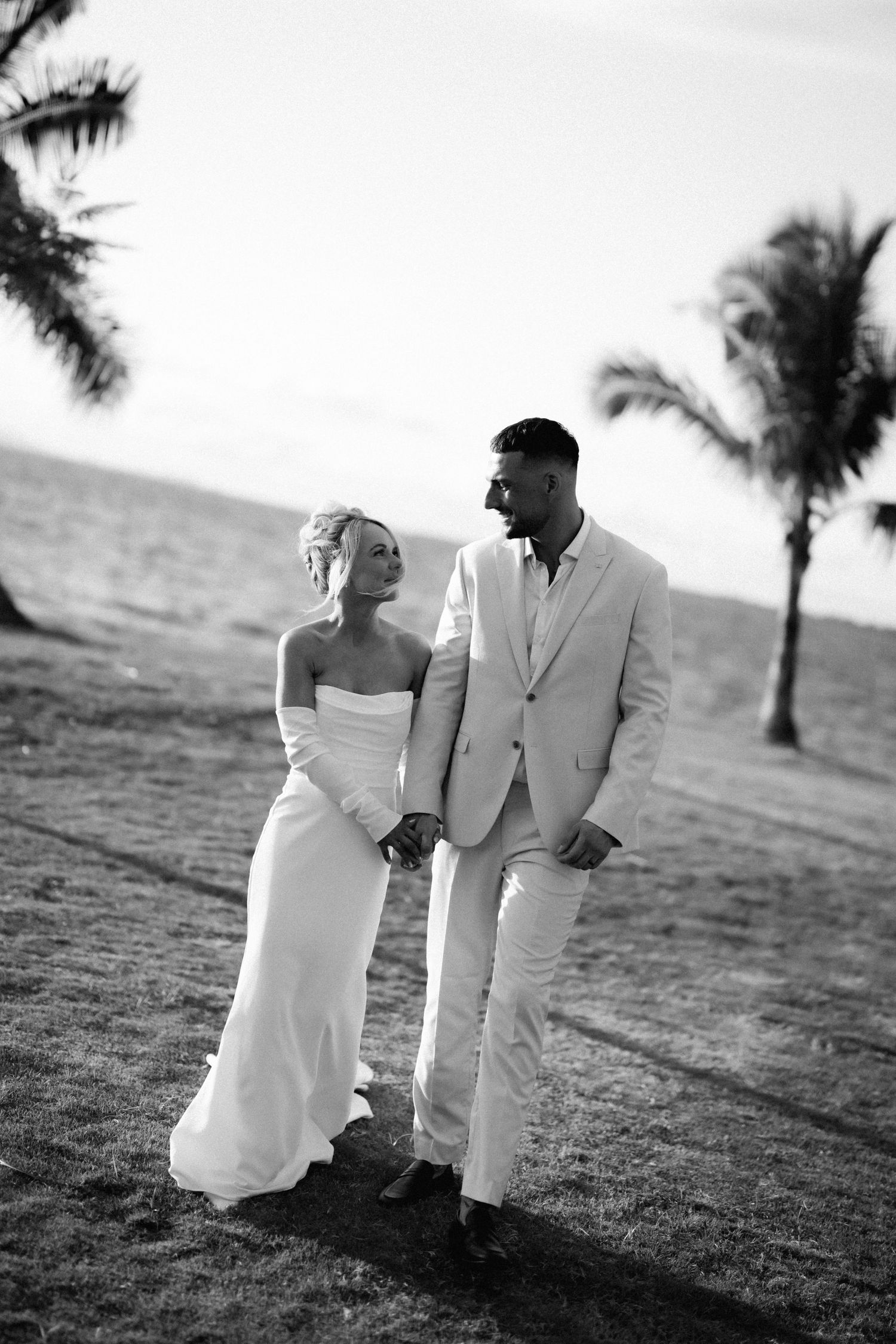 A romantic black and white photo of newlyweds walking together on a beach with palm trees in the background.