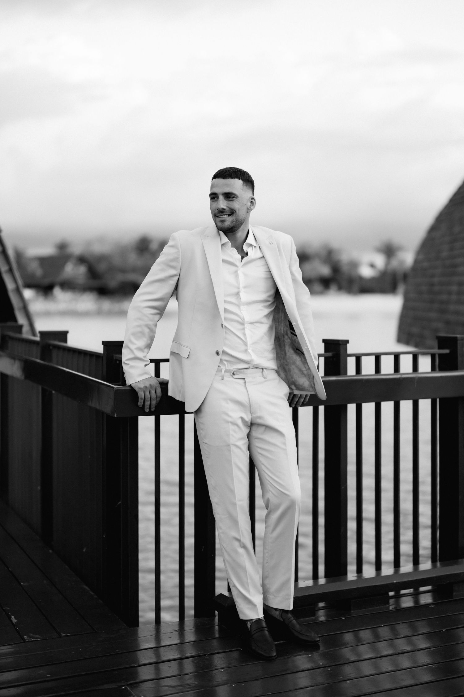 Groom in all-white suit leaning against deck railing with water view in background.
