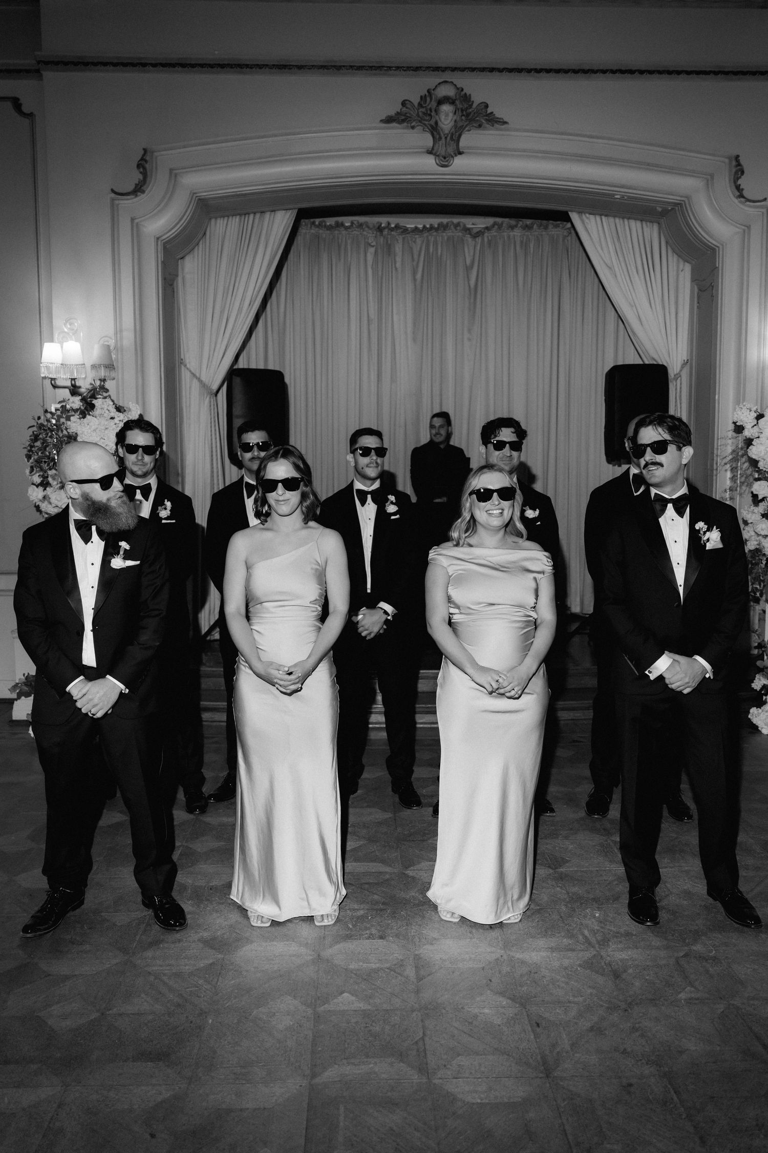 Wedding party wearing formal attire and sunglasses posing in front of white curtains.