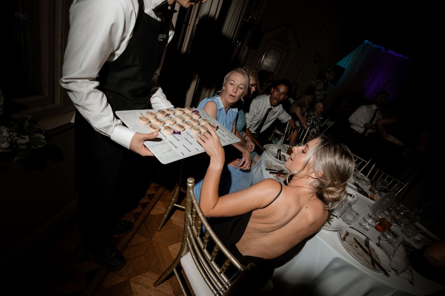 A waiter serves dessert to elegantly dressed guests at a wedding reception in a dimly lit venue.