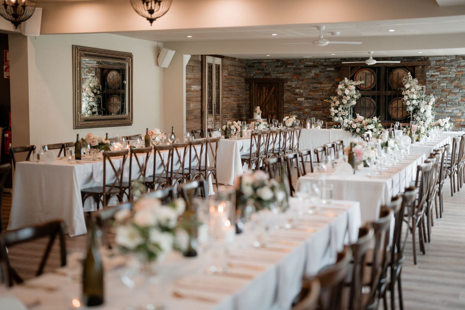 Elegant wedding reception venue with long tables, wooden cross-back chairs, and white floral centerpieces in a rustic room.