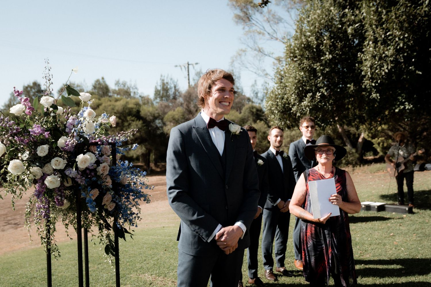 An outdoor wedding ceremony setup with a decorative floral arch and attendees gathered on a sunny day.