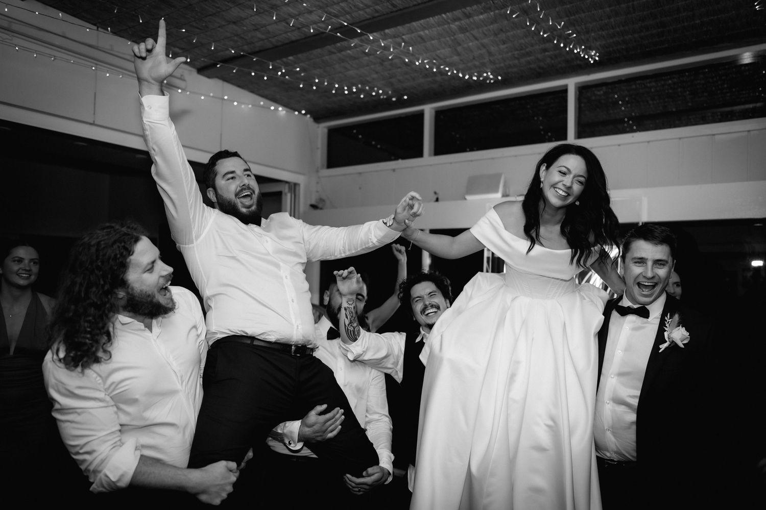Wedding party celebrates on dance floor with energetic moves and laughter in black and white photography.