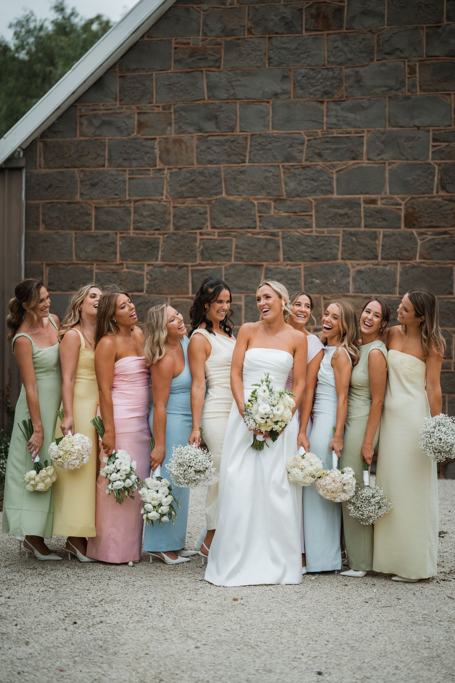 Wedding party lined up against stone wall wearing pastel colored gowns.