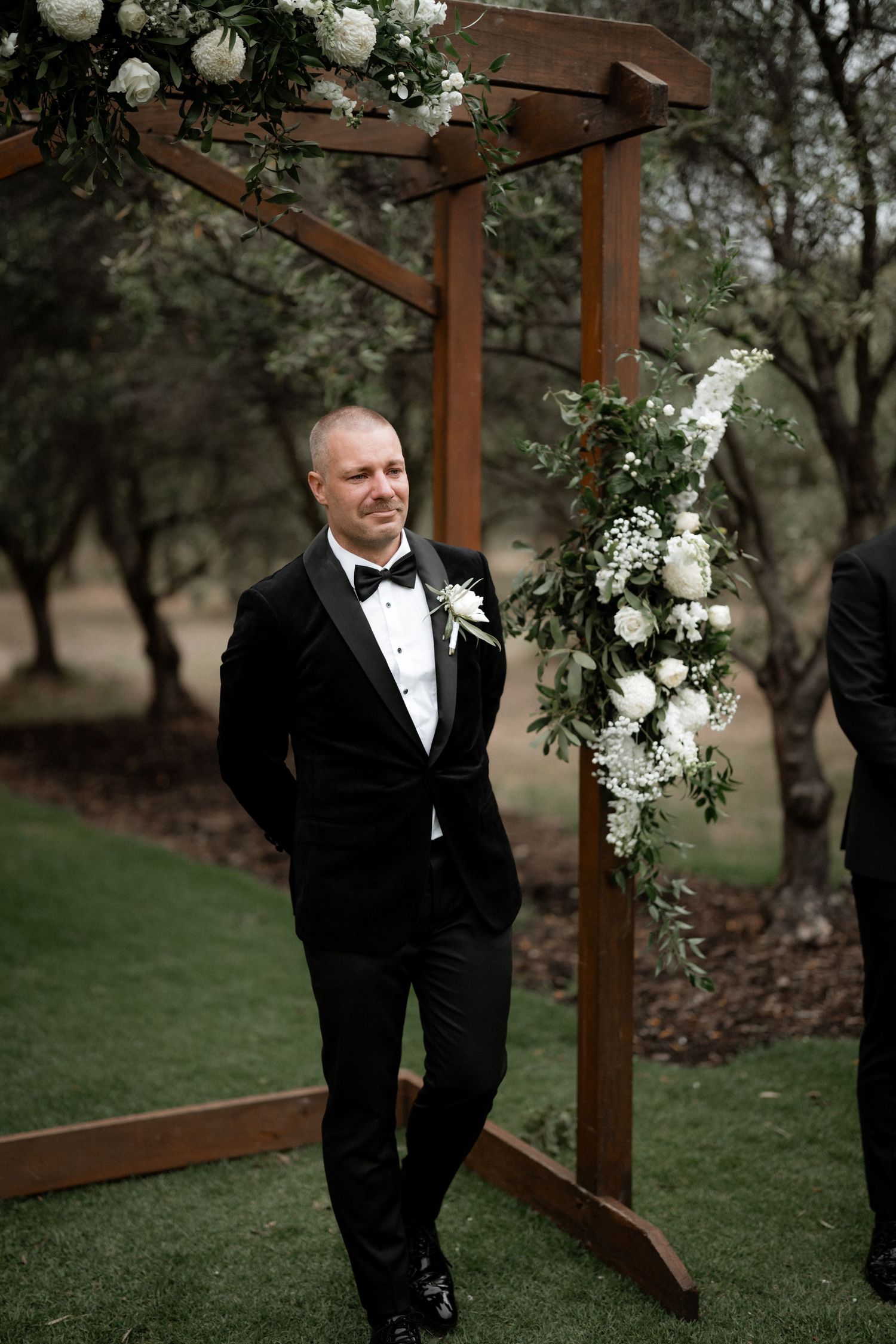 Groom in black tuxedo poses under wooden pergola arch decorated with white floral arrangements outdoors.