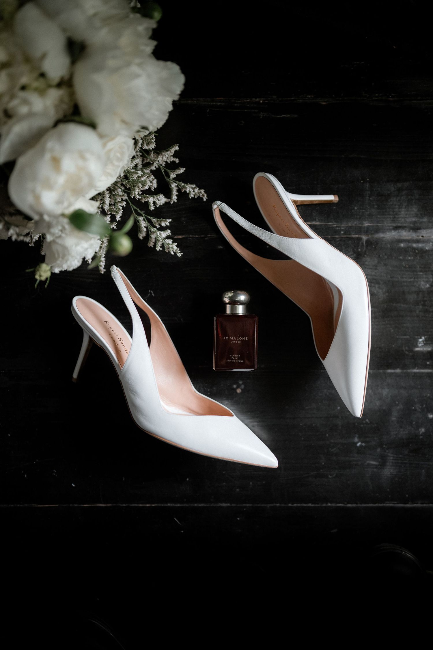 Elegant white slingback heels and perfume bottle arranged with white flowers on dark surface.