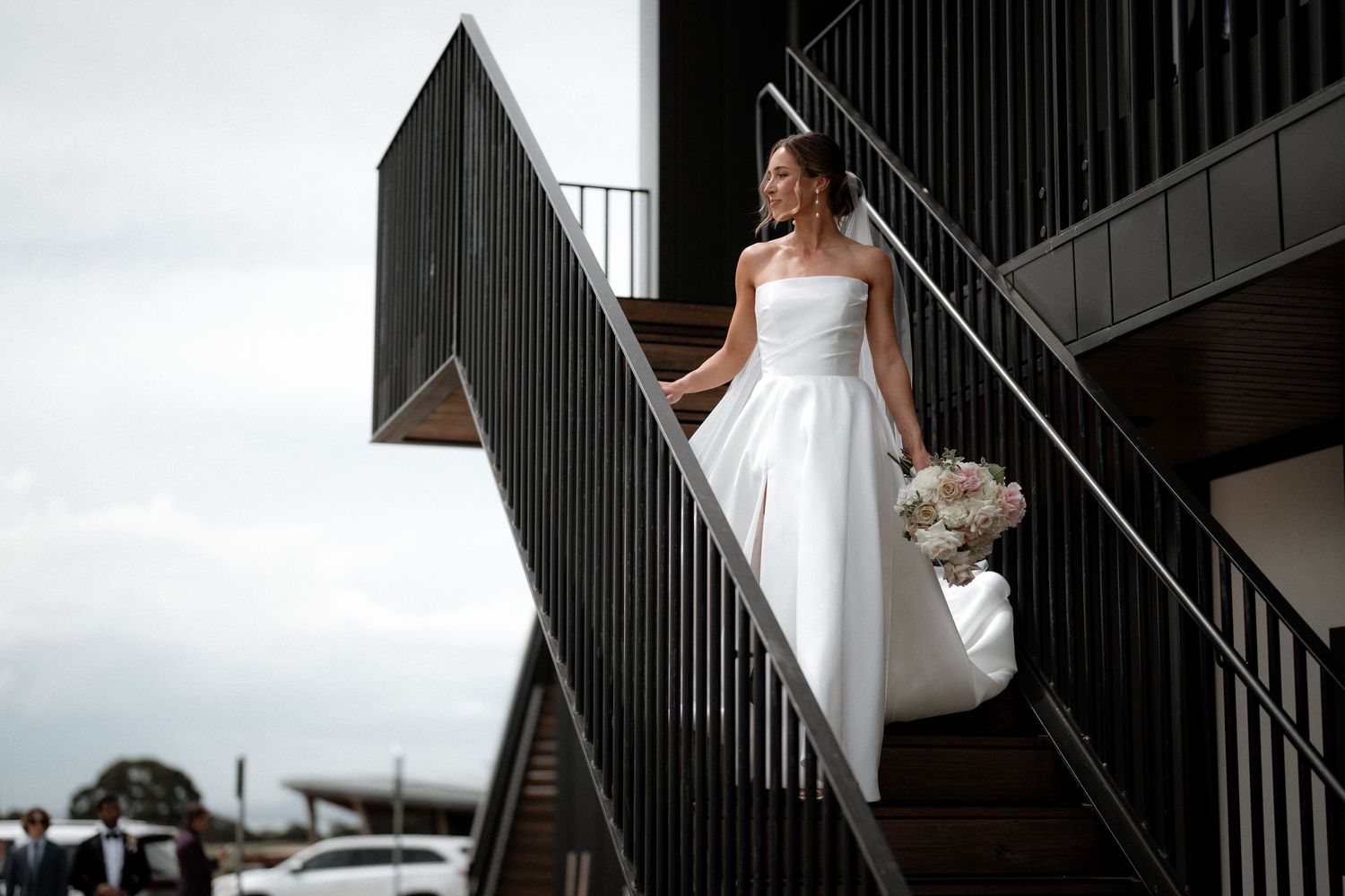 A bride in a white wedding dress walks down a modern staircase with black railings at a stylish venue.