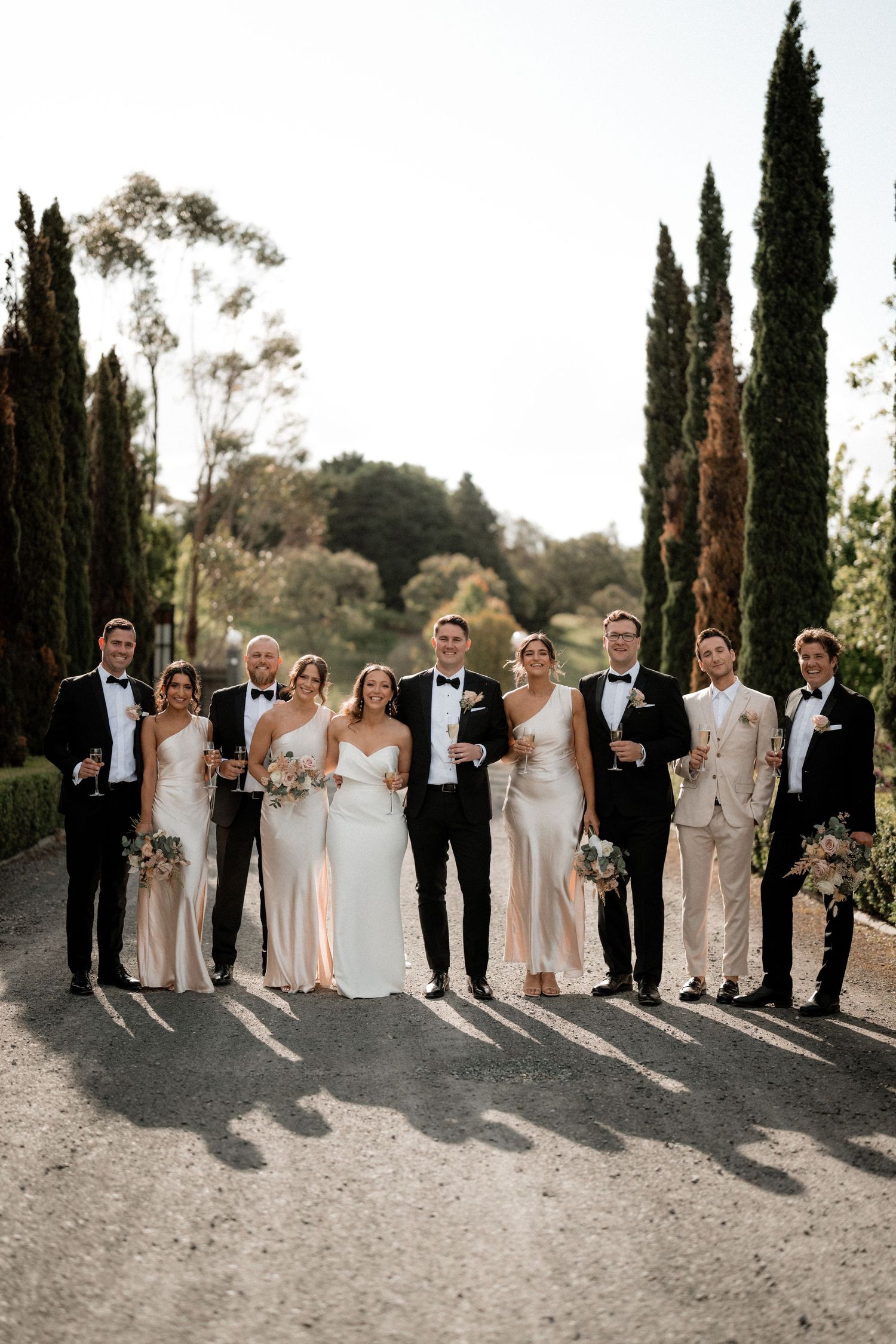 Wedding party poses outdoors in formal attire with cypress trees and natural setting in background during sunset.