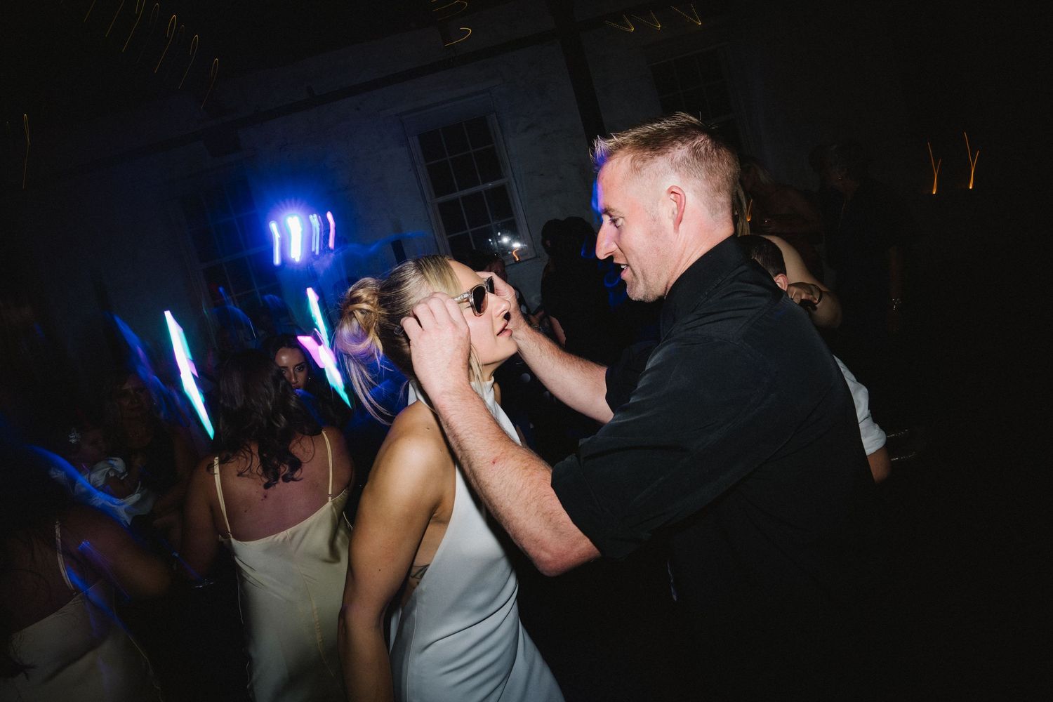 Two people dance close together under dark lighting with blue glow sticks visible in the background at a party.