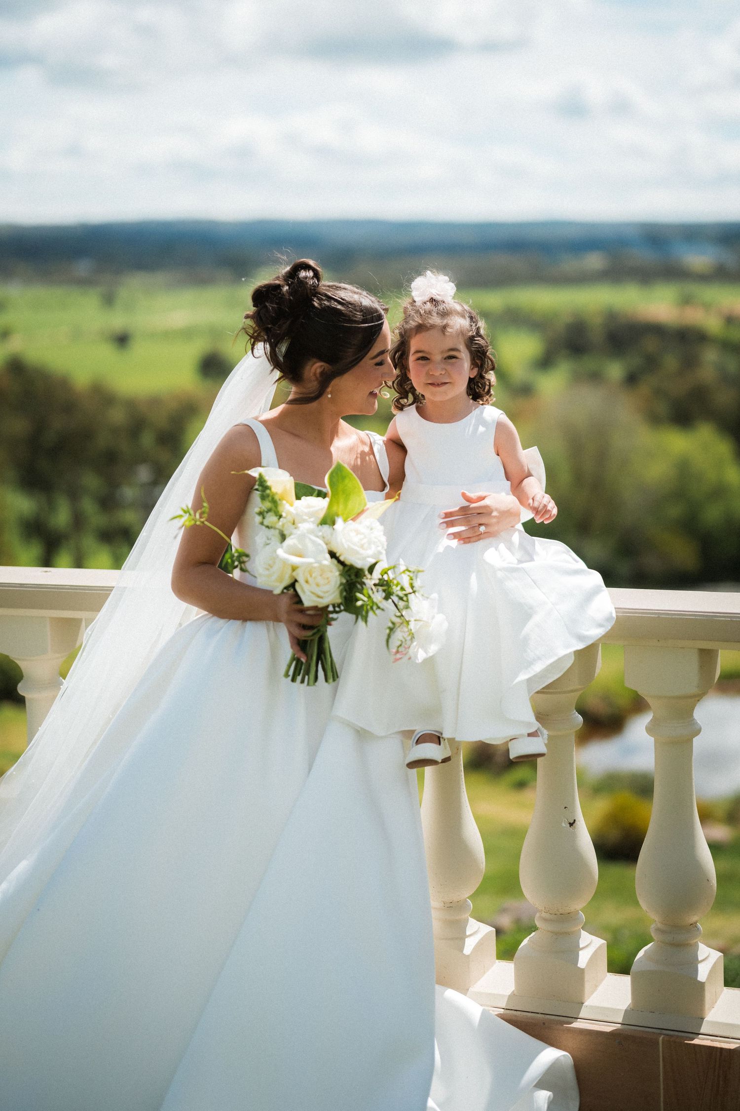 Bride in a white gown holding a white bouquet sits with her flower girl on a stone balustrade overlooking green countryside.