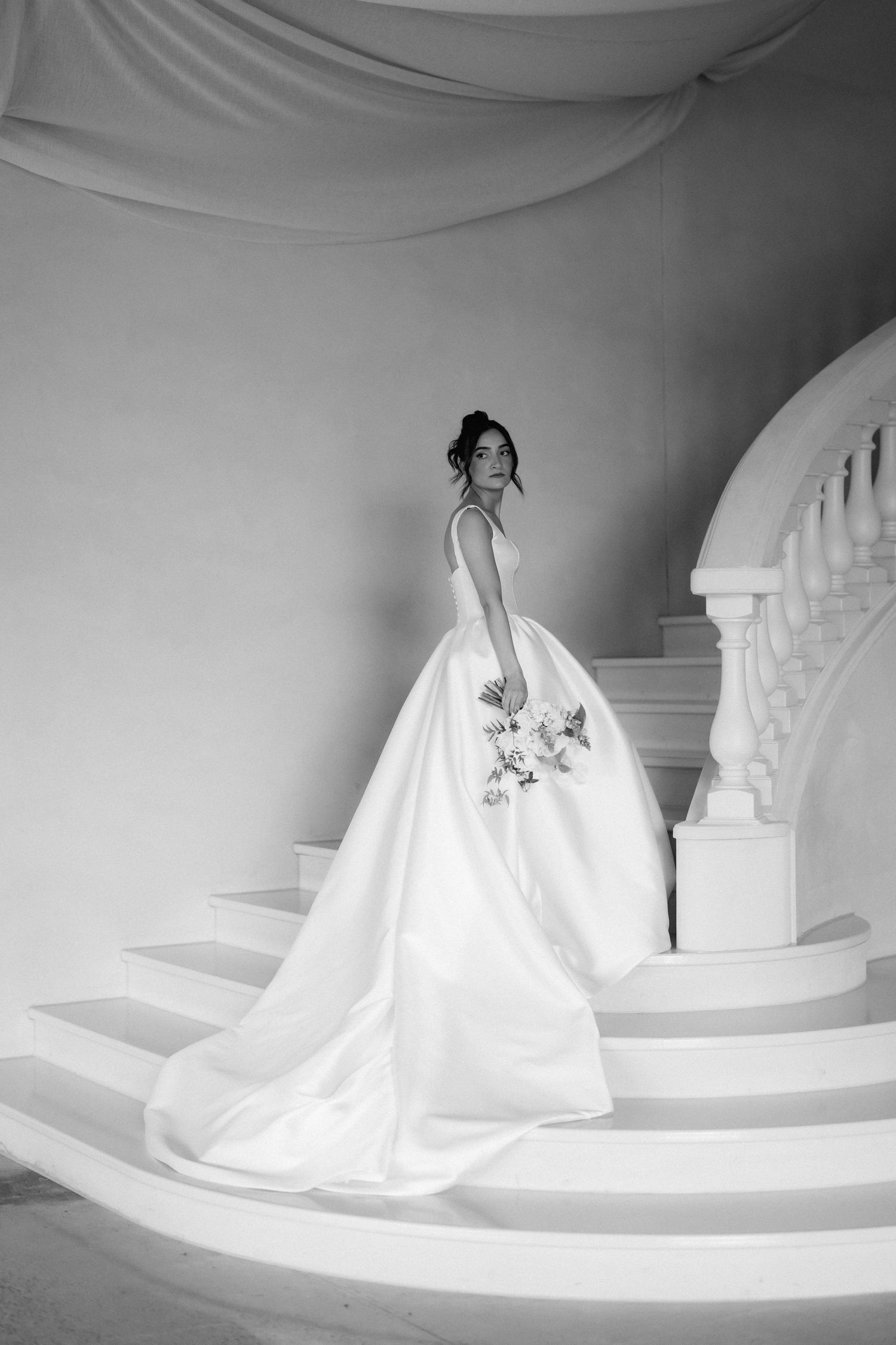Black and white photo of a bride in an elegant satin ball gown holding a bouquet while standing on a grand curved staircase.