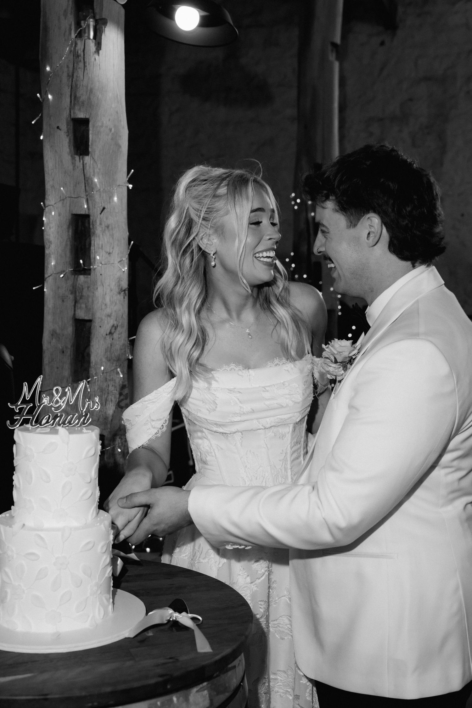 Black and white photo of bride and groom laughing while cutting white wedding cake together.