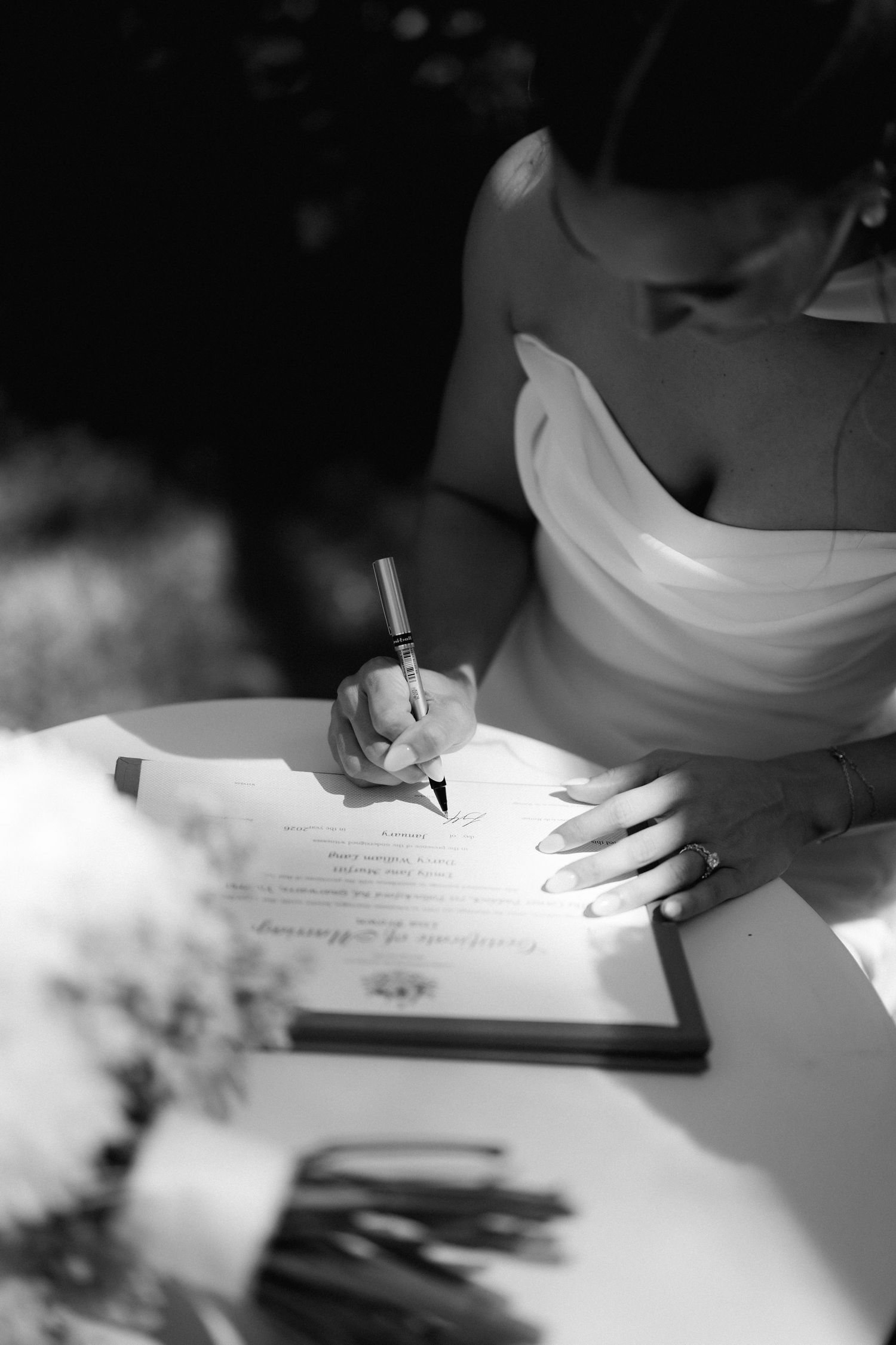 Close-up black and white shot of hands writing in a wedding guest book or signing documents.