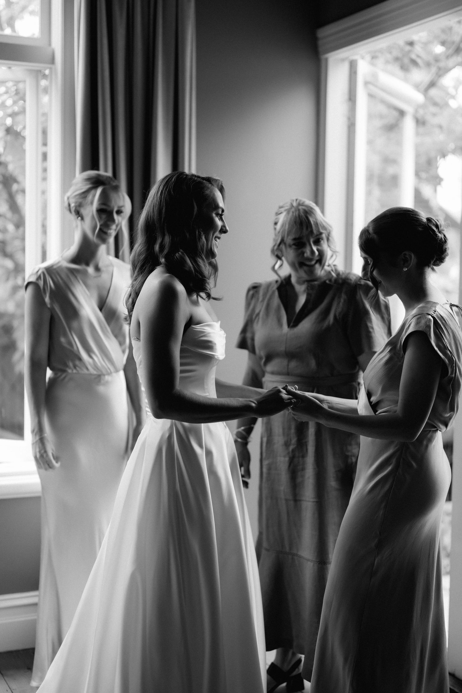 Bridesmaids in matching gray dresses stand by sunlit windows during wedding preparations.