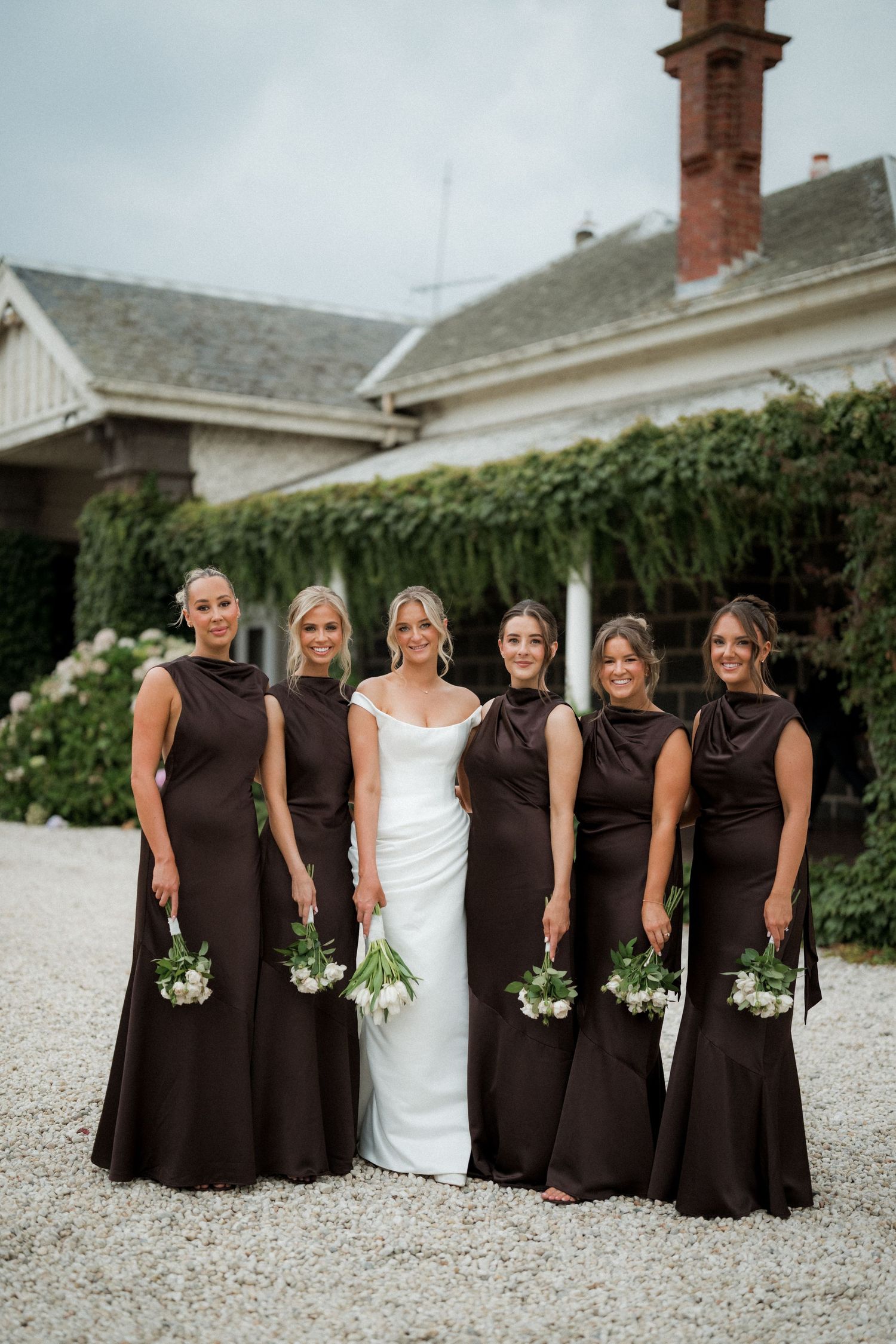 Bridesmaids in matching brown dresses pose with the bride in white on a gravel path.