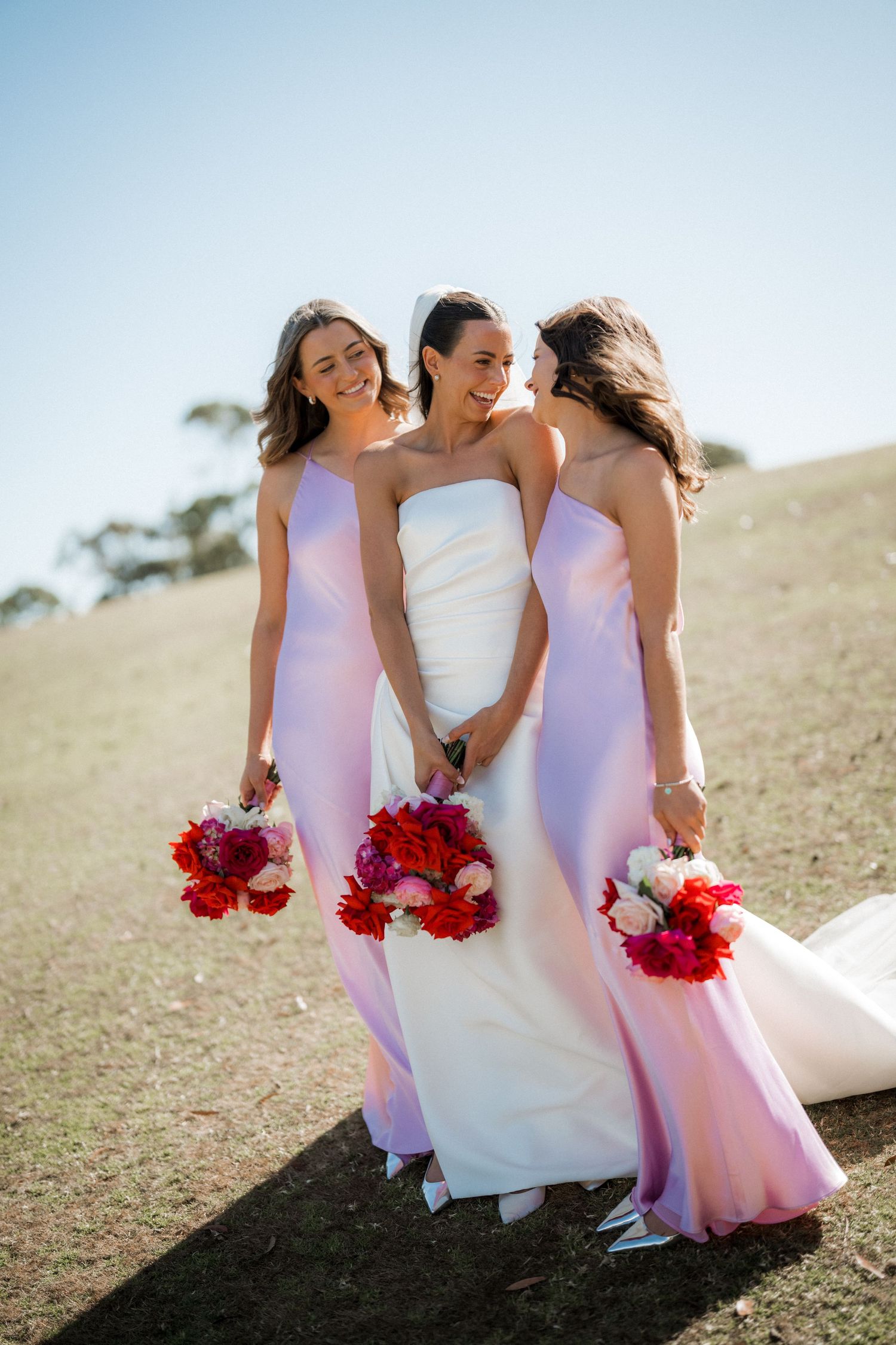 Bridesmaids in pink dresses and bride in white holding vibrant red bouquets pose together on grassy hillside.