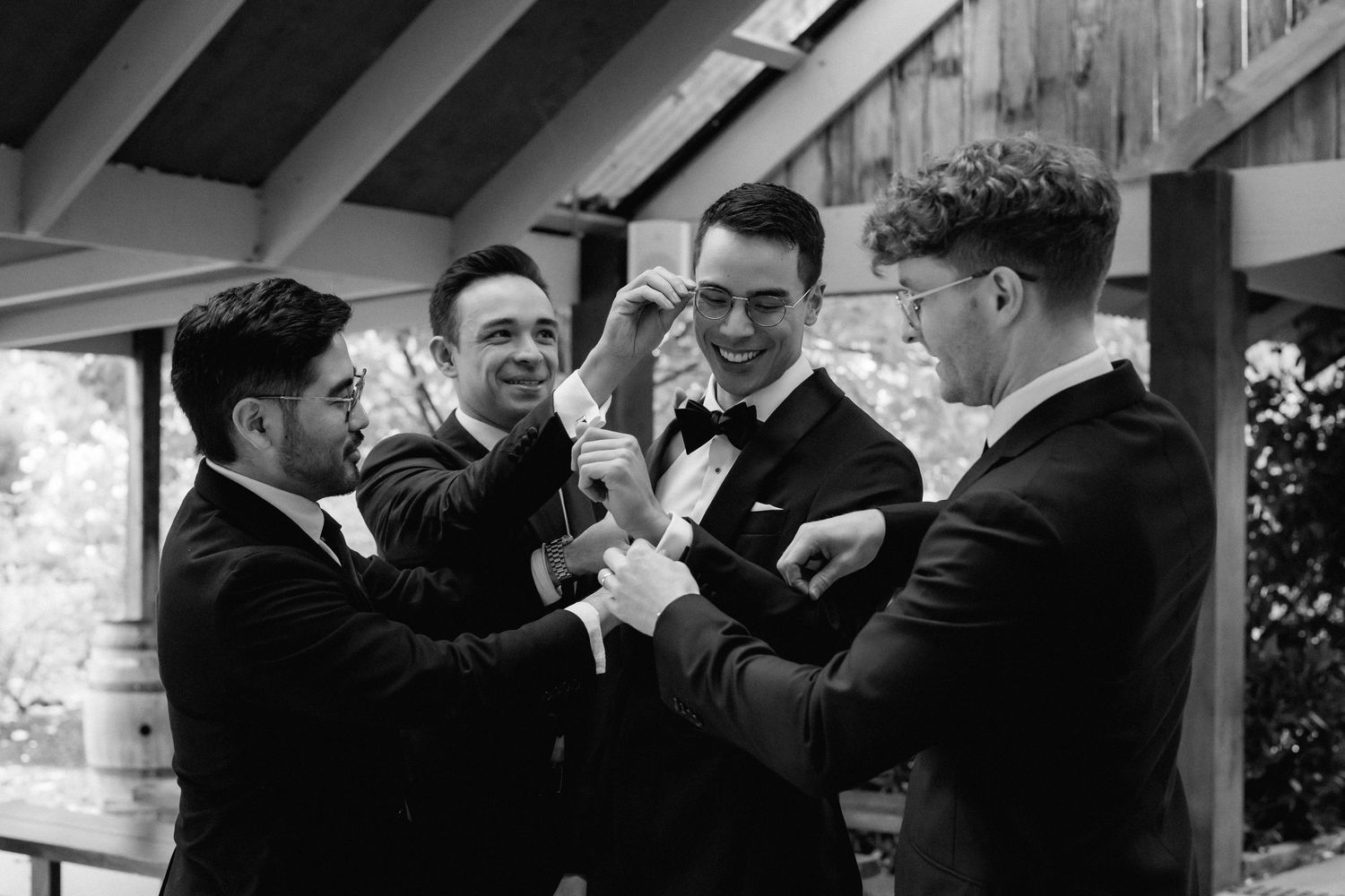 Groomsmen in black tuxedos helping groom with cufflinks before wedding ceremony, black and white photo.