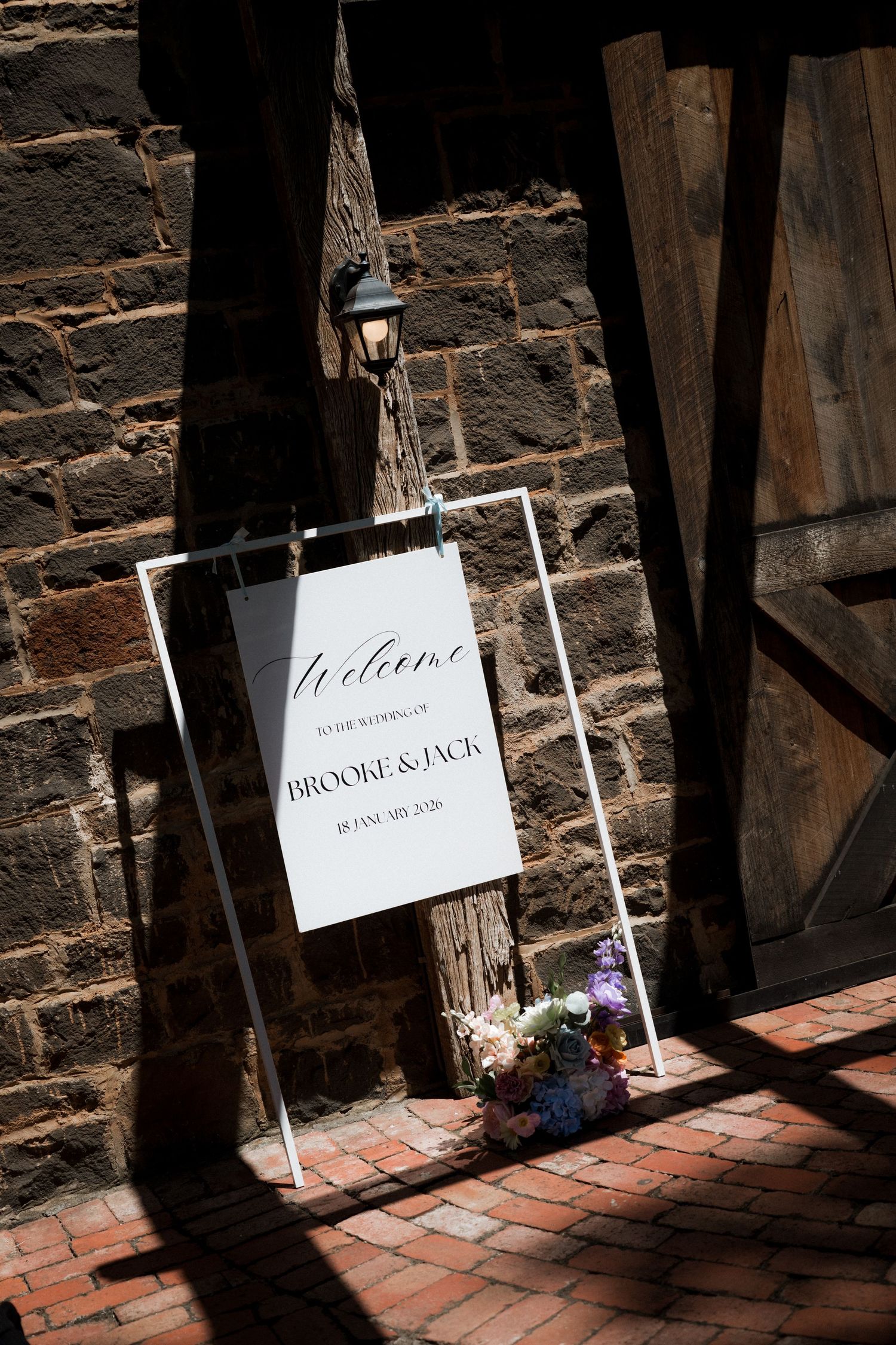Welcome sign for Brooke and Jack wedding leaning against rustic brick wall in dappled sunlight.