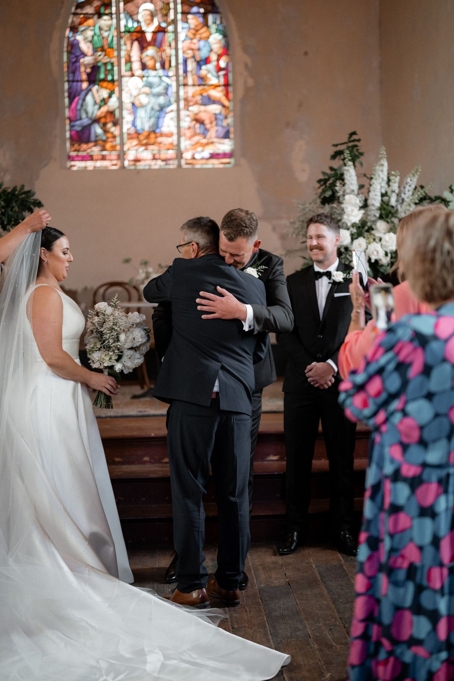 An emotional moment during a wedding ceremony inside a chapel with stained glass windows and floral decorations.