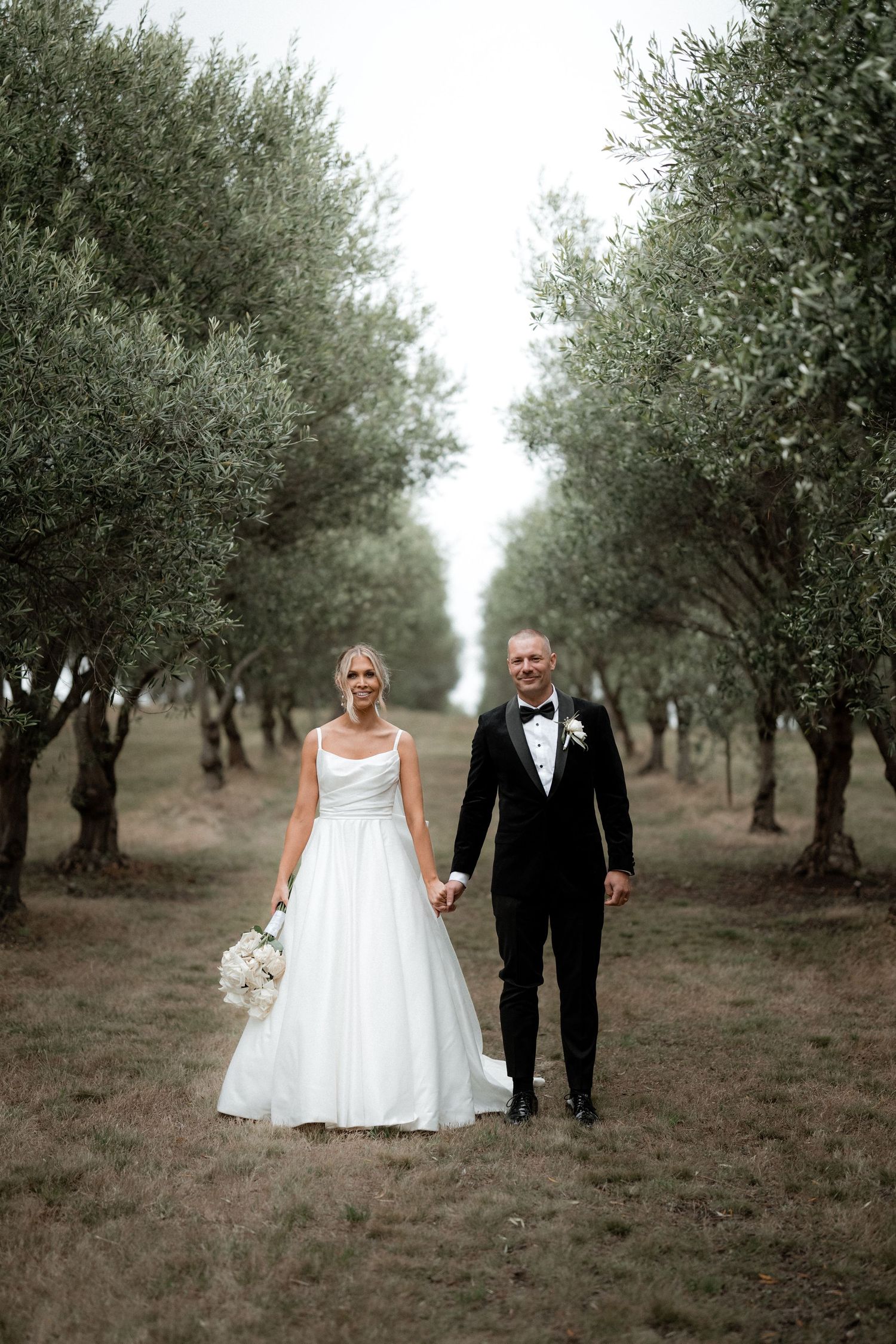 A bride and groom pose for romantic wedding photos in an olive grove pathway.