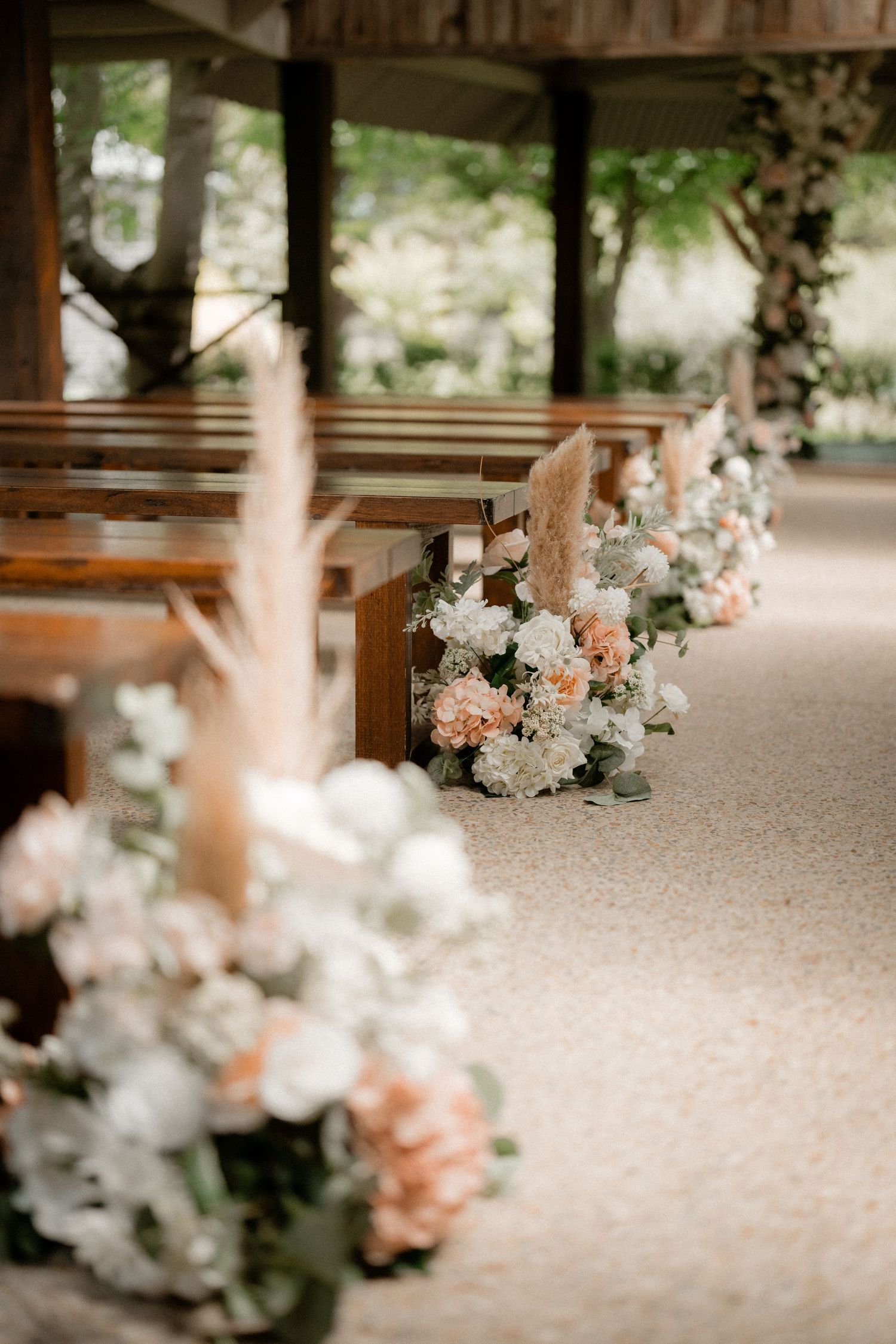 Elegant flower arrangements with white and peach blooms decorate wooden pews in a rustic wedding ceremony venue.