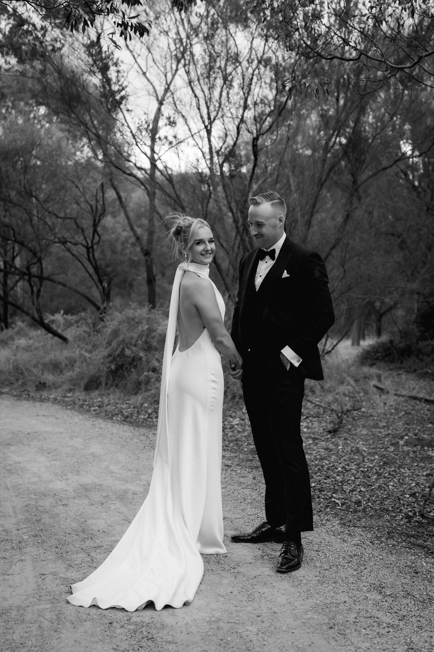 Black and white wedding photo of a couple on a forest path, bride in backless dress with long train, groom in tuxedo.