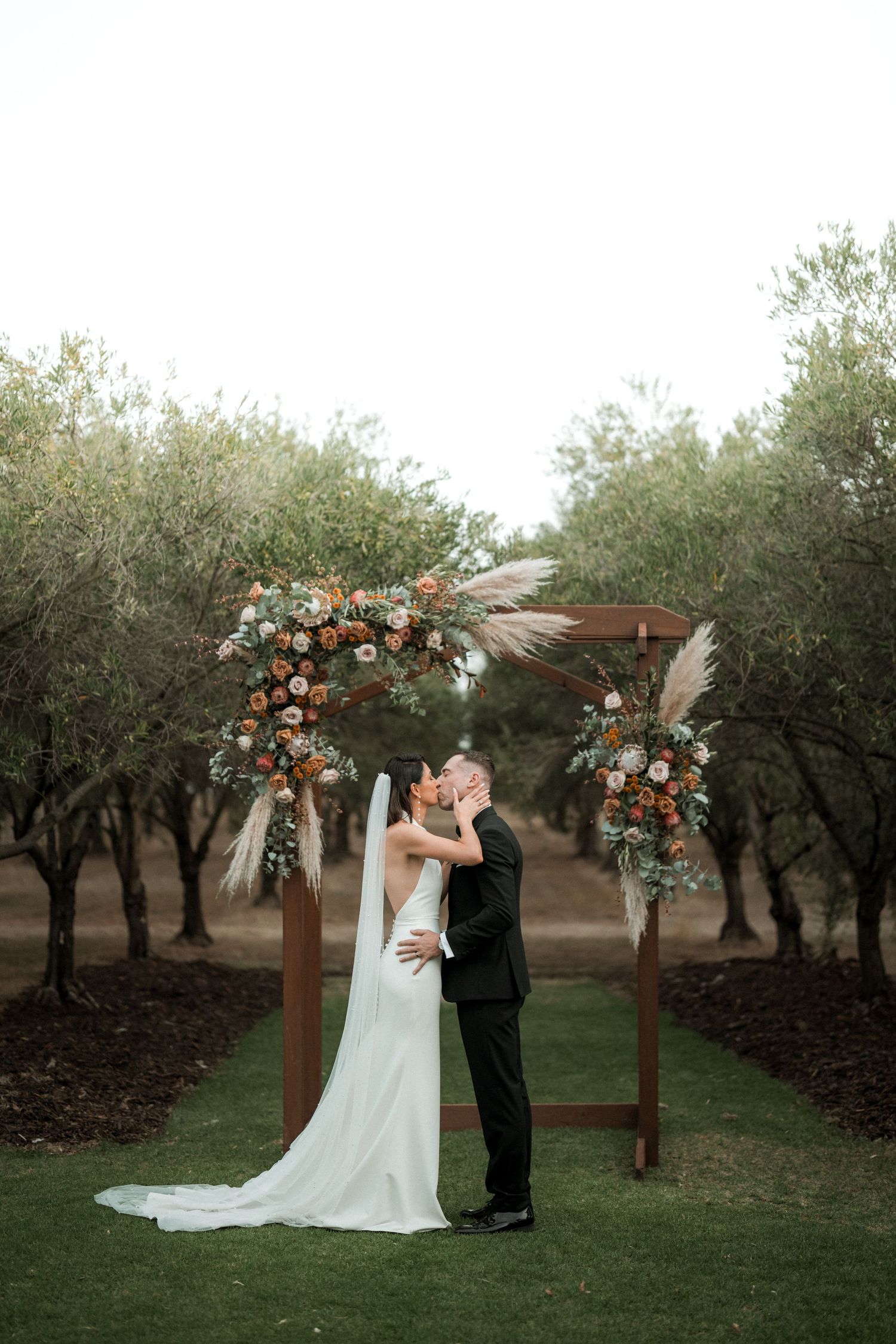 Newlyweds share a romantic moment under a wooden arch adorned with peach and white floral arrangements in an olive grove.
