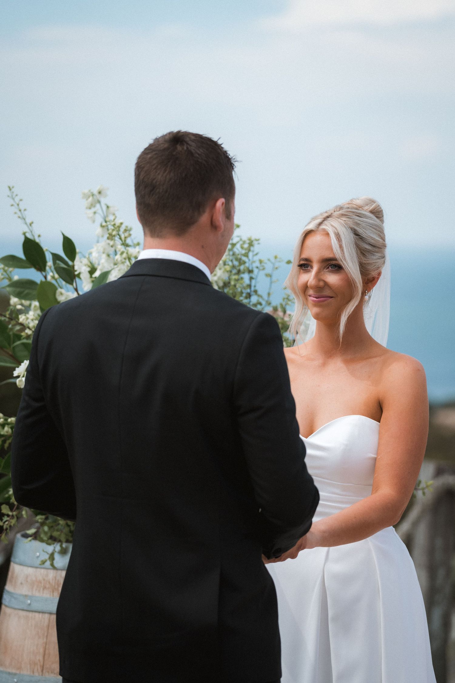 Wedding ceremony moment with white floral arrangements and rustic barrels against an ocean backdrop.