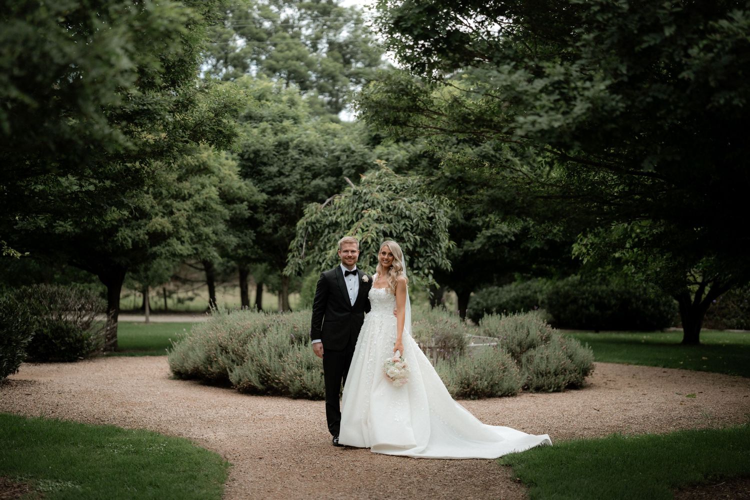 Elegant wedding couple poses on gravel path surrounded by manicured garden landscaping.