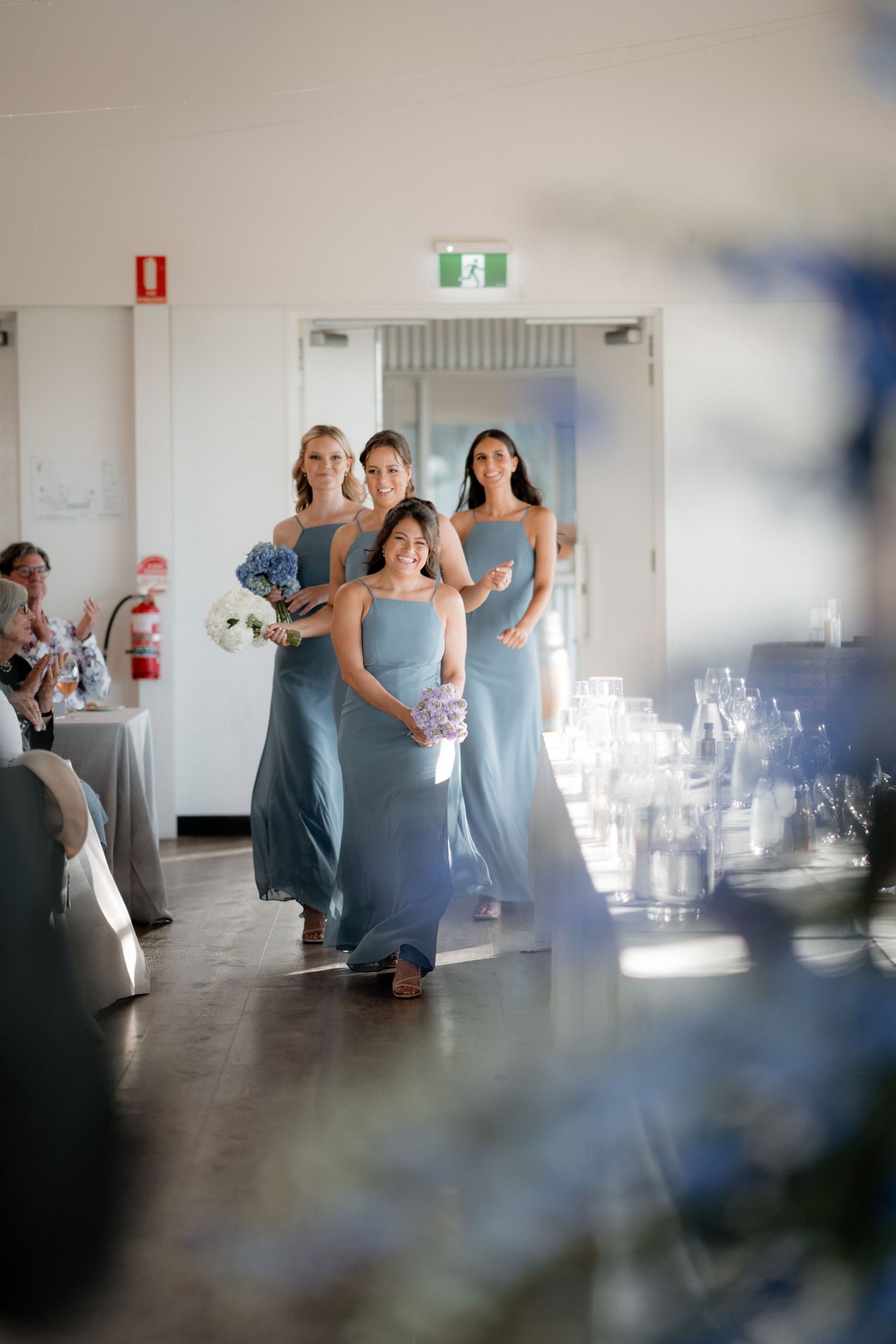 Bridesmaids in flowing light blue dresses walk gracefully through an indoor venue decorated with glowing candles.