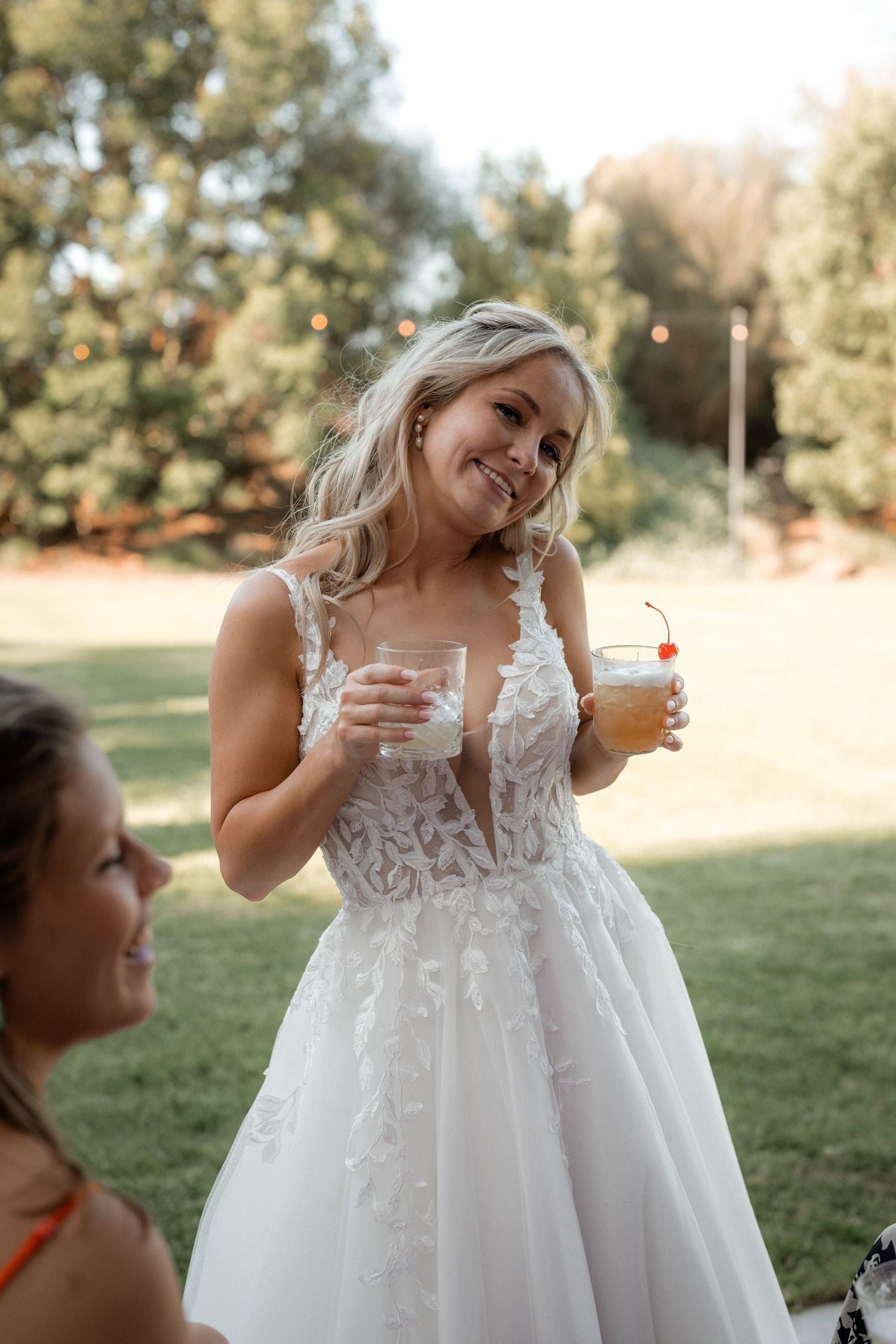 Bride in lace wedding dress smiling while holding cocktails outdoors.