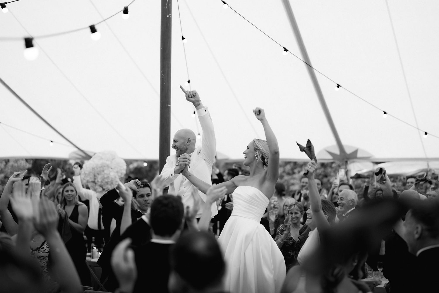Dancing couple in wedding attire surrounded by cheering crowd under festive string lights at outdoor reception.