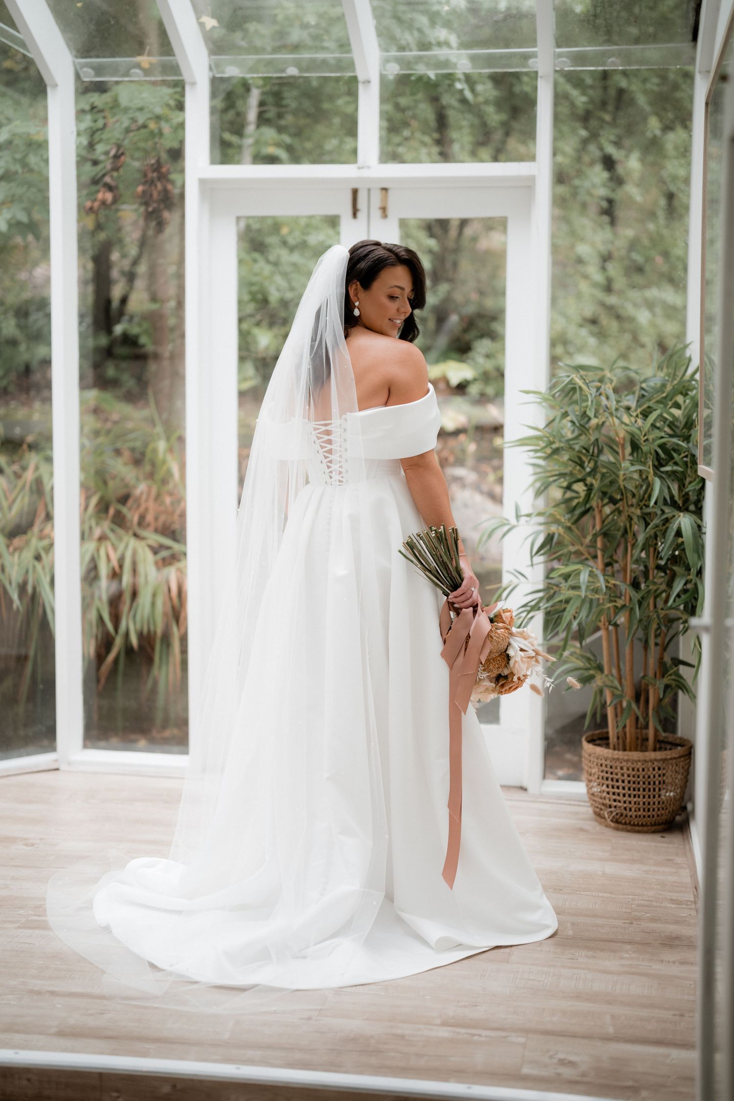 A bride in an elegant off-shoulder white wedding gown and long veil holds a bouquet in a sunlit conservatory.