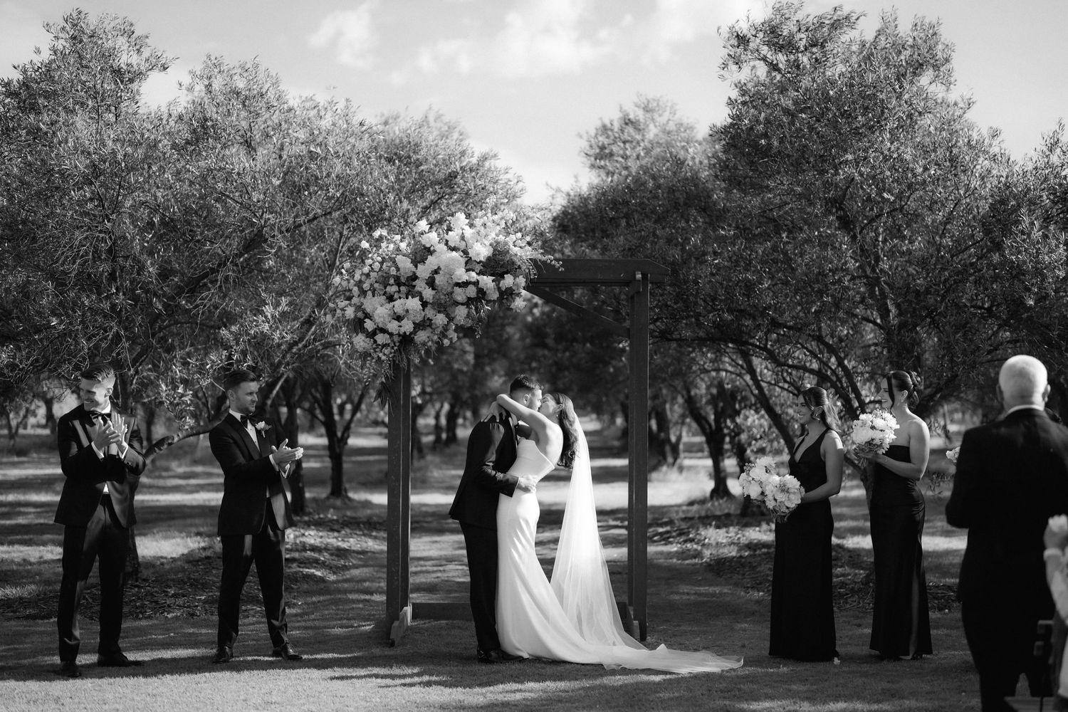 A black and white wedding ceremony takes place in a serene orchard setting with blooming trees in the background.