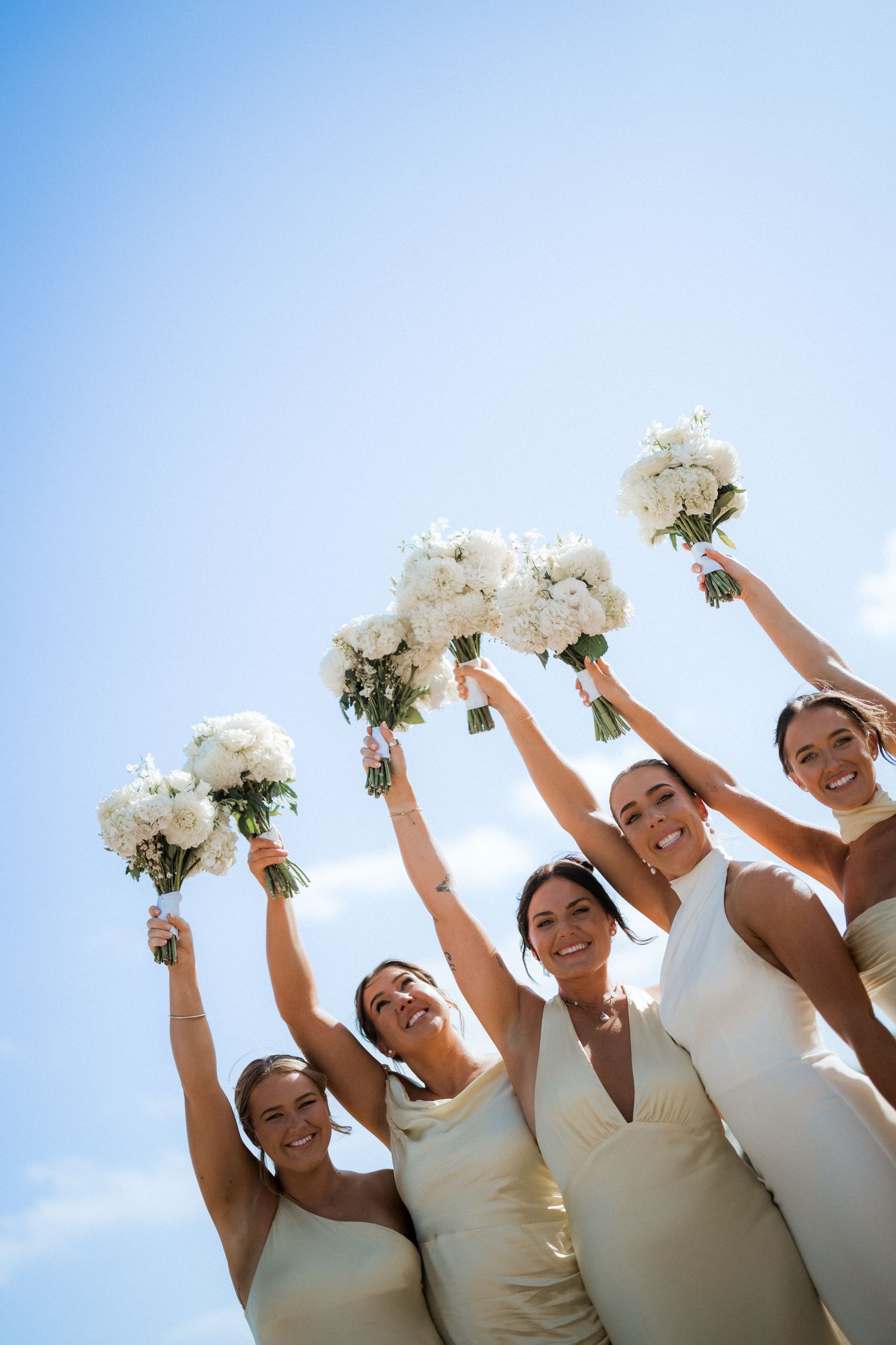 Bridesmaids in cream dresses holding white bouquets against blue sky.