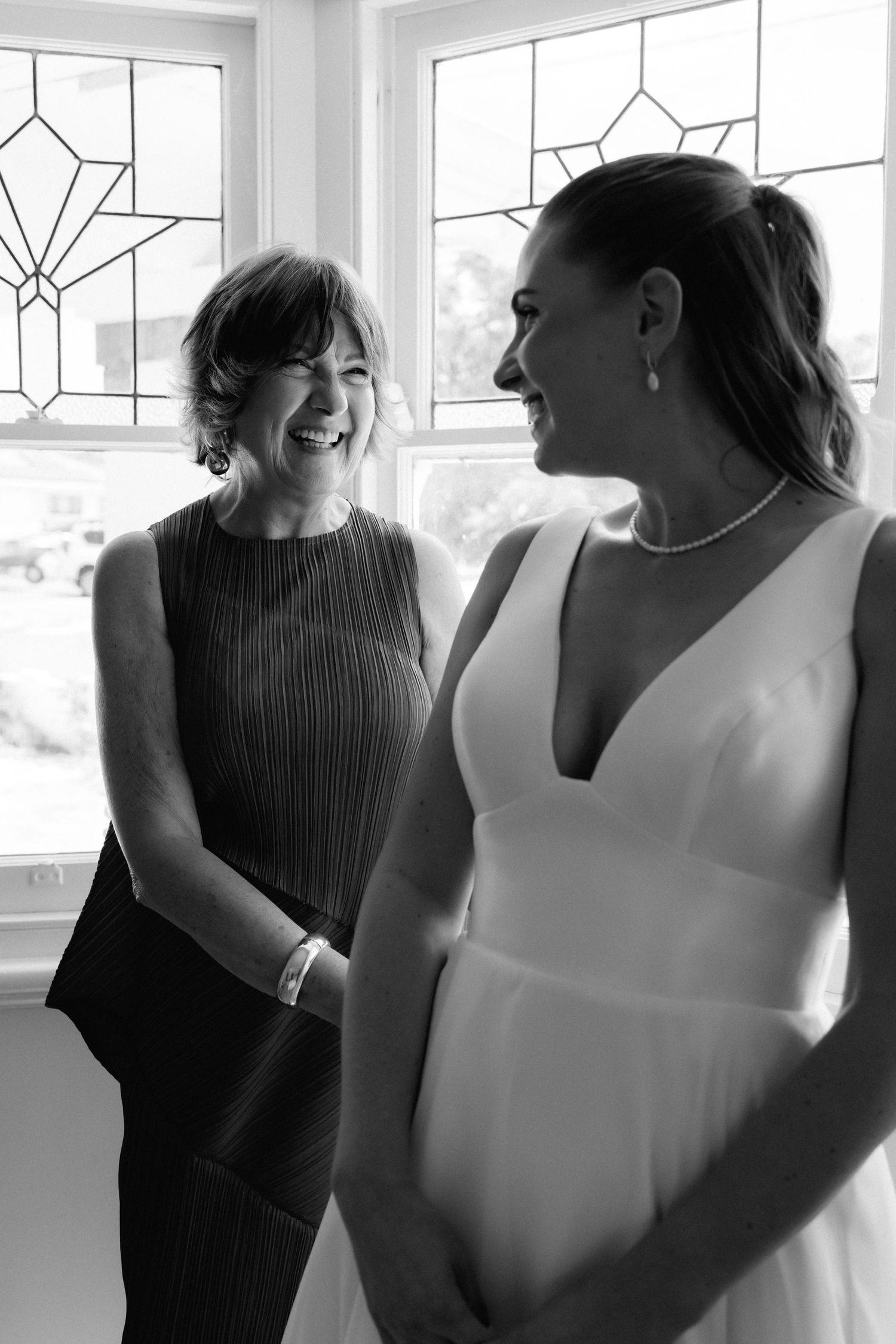 Two wedding attendants share a joyful moment in front of decorative windows.