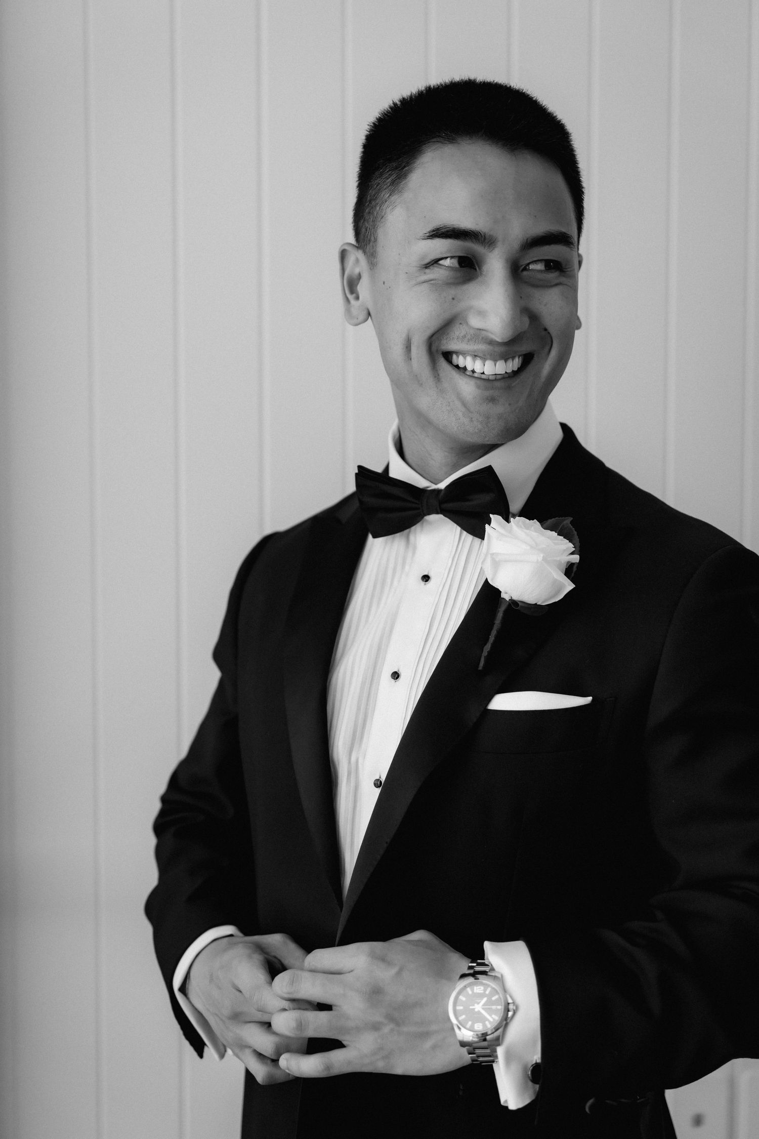 A formal portrait of a groom in a classic black tuxedo with bow tie and white boutonniere smiling at the camera.